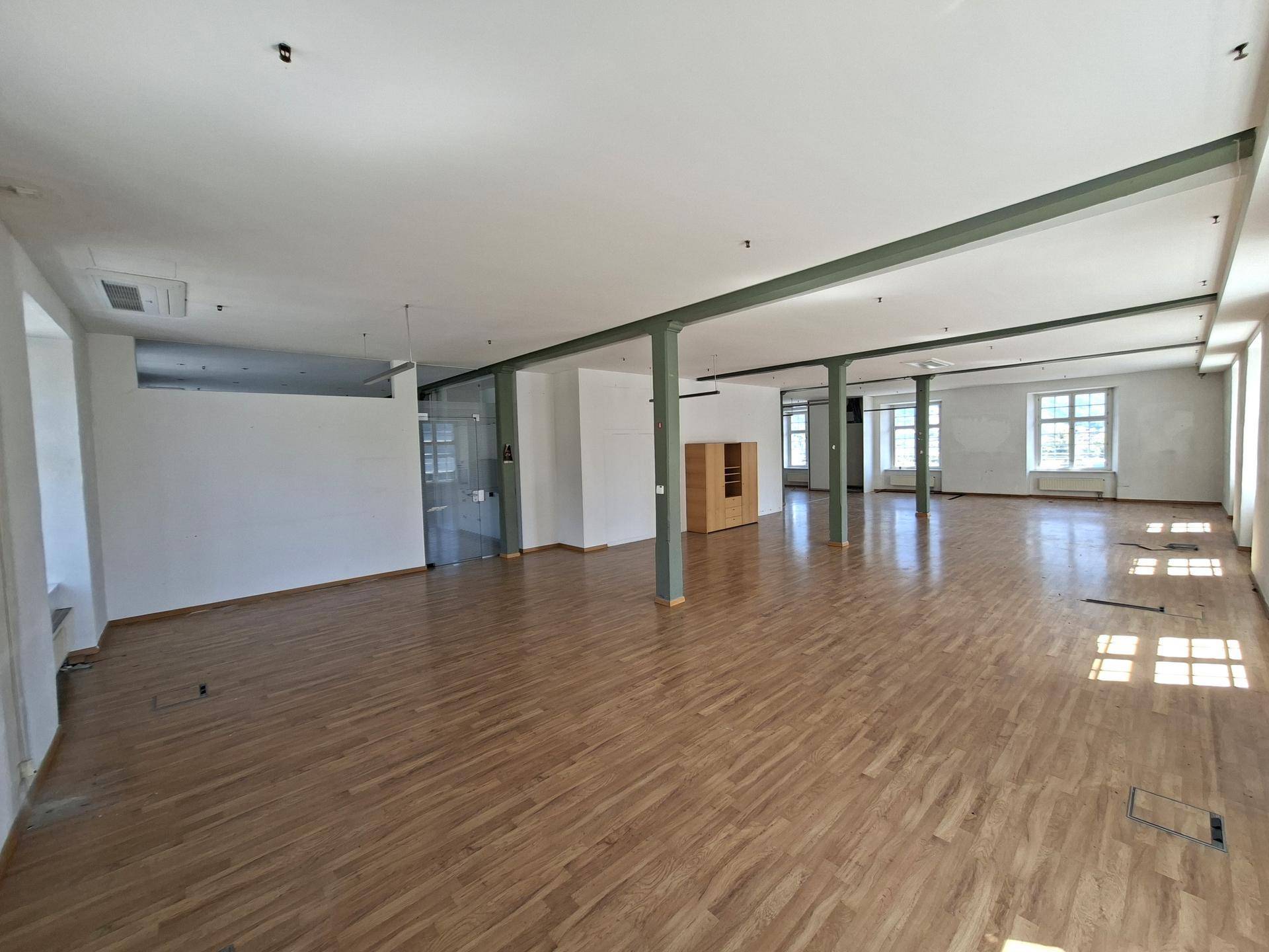 Weitläufiger offener Bürobereich mit Holzboden, grünen Trägern und einer Glasabtrennung.