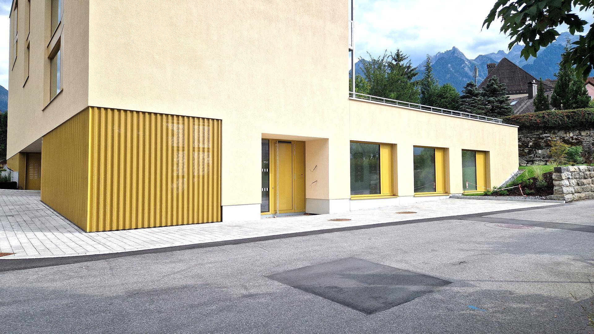 Eingangsbereich des modernen Gebäudes mit beiger Fassade, Holzdetails und großen Fenstern zur Straße hin.