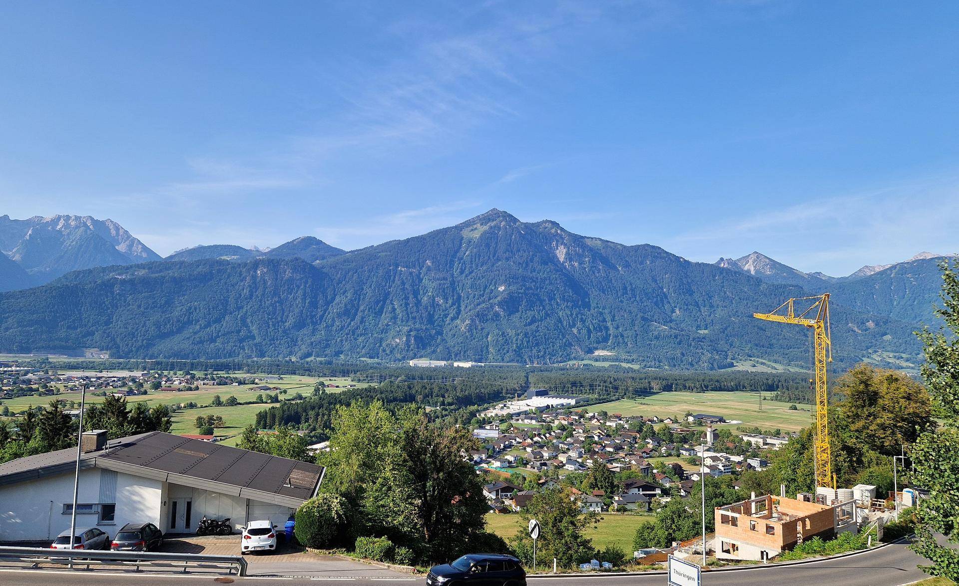 Panoramablick auf ein Tal mit einer Stadt, umrahmt von Bergen, inklusive einer Baustelle mit Kran.