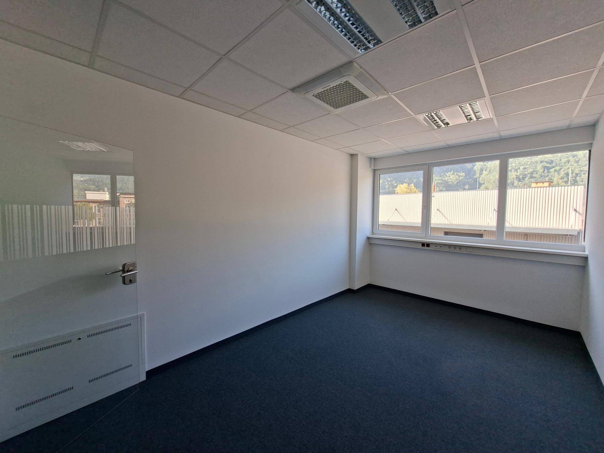Helles Büro mit Teppichboden, großen Fenstern und einer modernen Glastür.