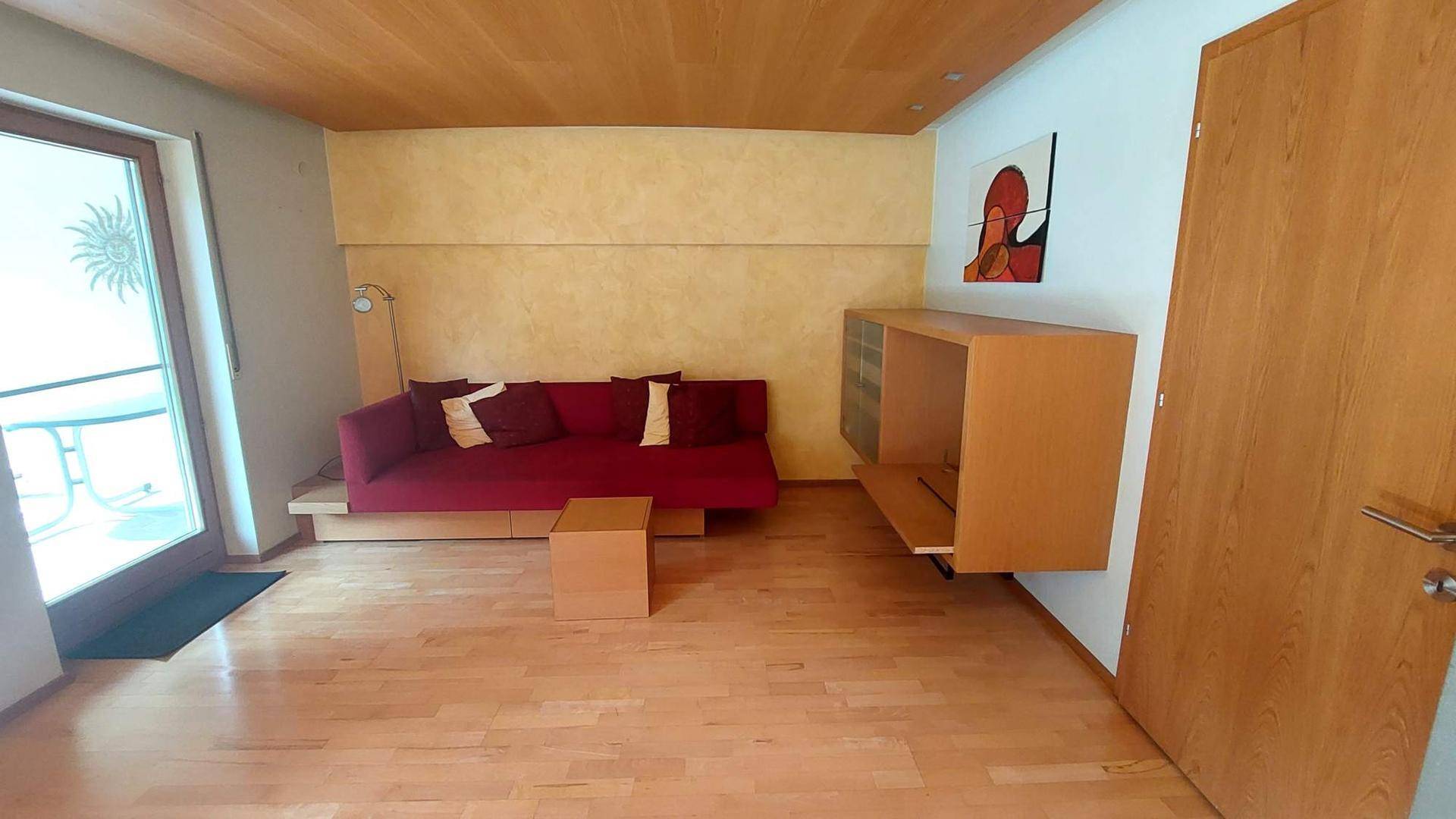 Wohnzimmer mit Holzboden, rotem Sofa und Zugang zu einem Außenbereich, unter einer Holzdecke.