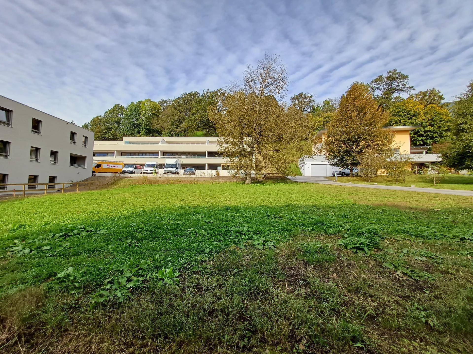 Weitläufige Grünfläche mit Blick auf eine moderne Wohnanlage und bewaldete Hügel im Hintergrund.
