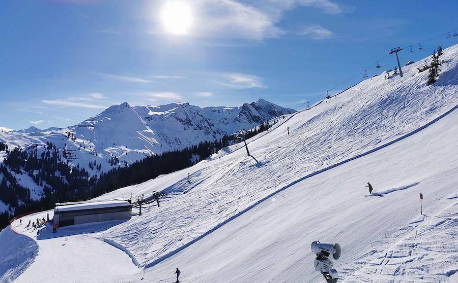 Sonnige Skipiste mit Skifahrern und beeindruckendem Alpenpanorama im Hintergrund.
