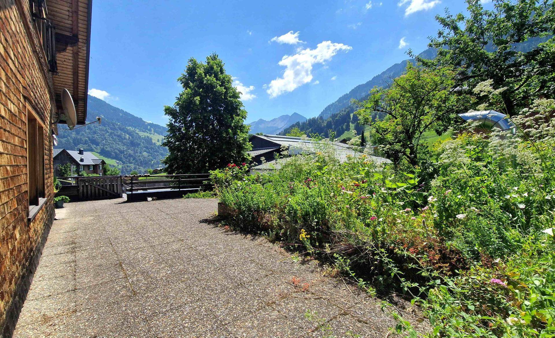 Außenbereich mit gepflastertem Weg, Blumenbeeten und Bergblick.