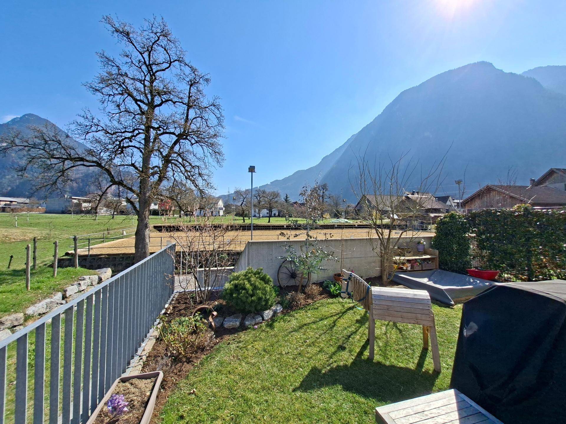 Sonniger Gartenbereich mit Rasenfläche, Pflanzen und weitem Blick auf die umliegende Berglandschaft.