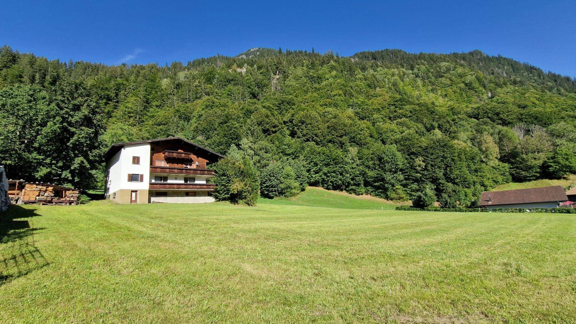 Alpines Wohnhaus mit traditionellen Holzbalkonen vor einer bewaldeten Bergkulisse und weitem Grün.