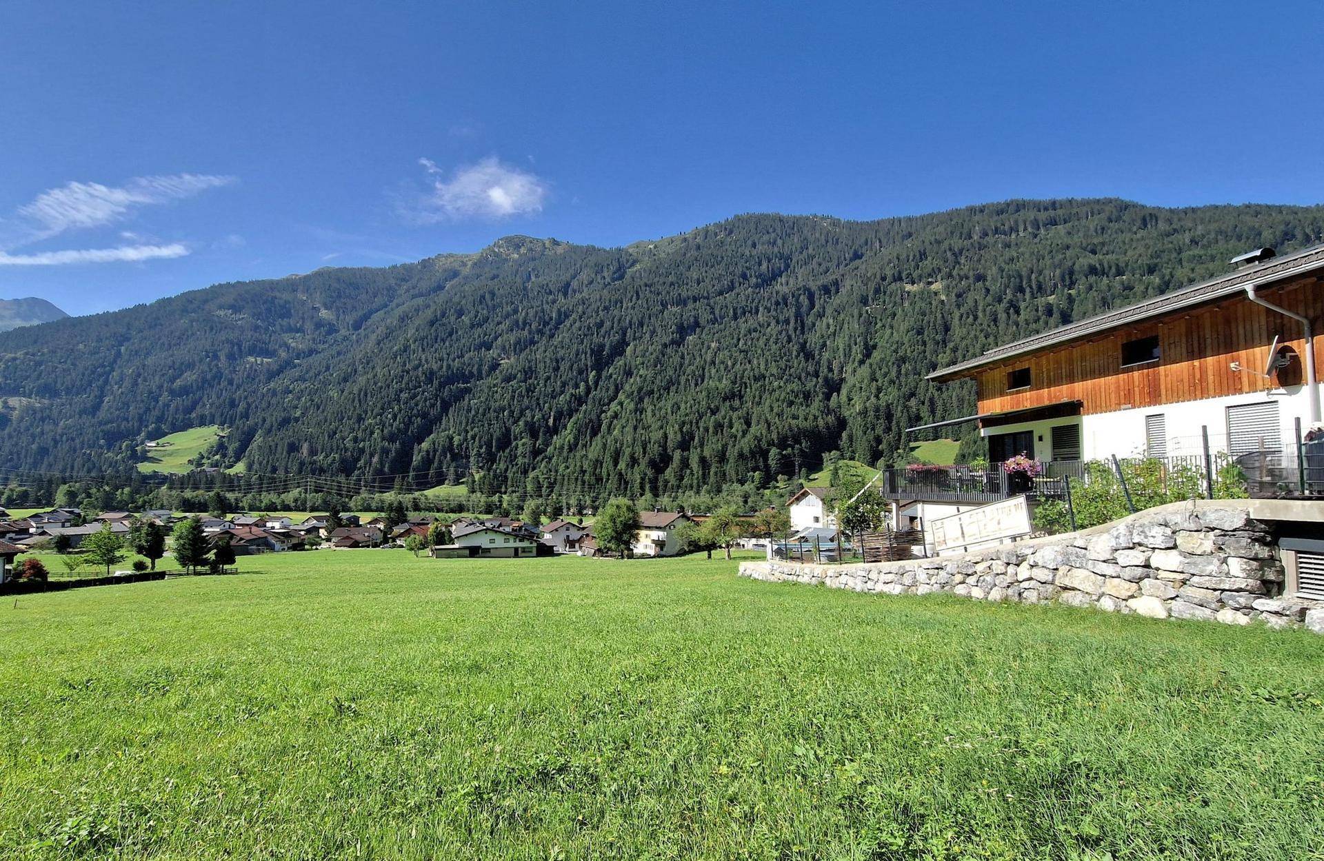 Weitläufiger Blick über die grüne Landschaft und ein Dorf mit Bergpanorama im Hintergrund.