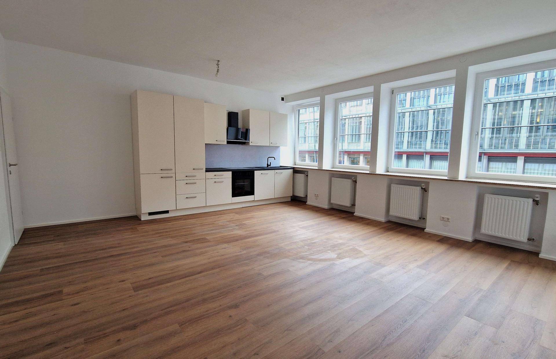Offener Wohnbereich mit integrierter Küchenzeile, Laminatboden und großen Fenstern, die viel Licht hereinlassen.