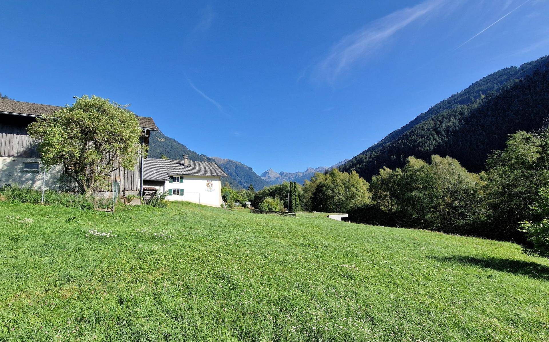 Häuser eingebettet in eine weite grüne Wiesenlandschaft mit majestätischen Bergen am Horizont, sonniger Tag.