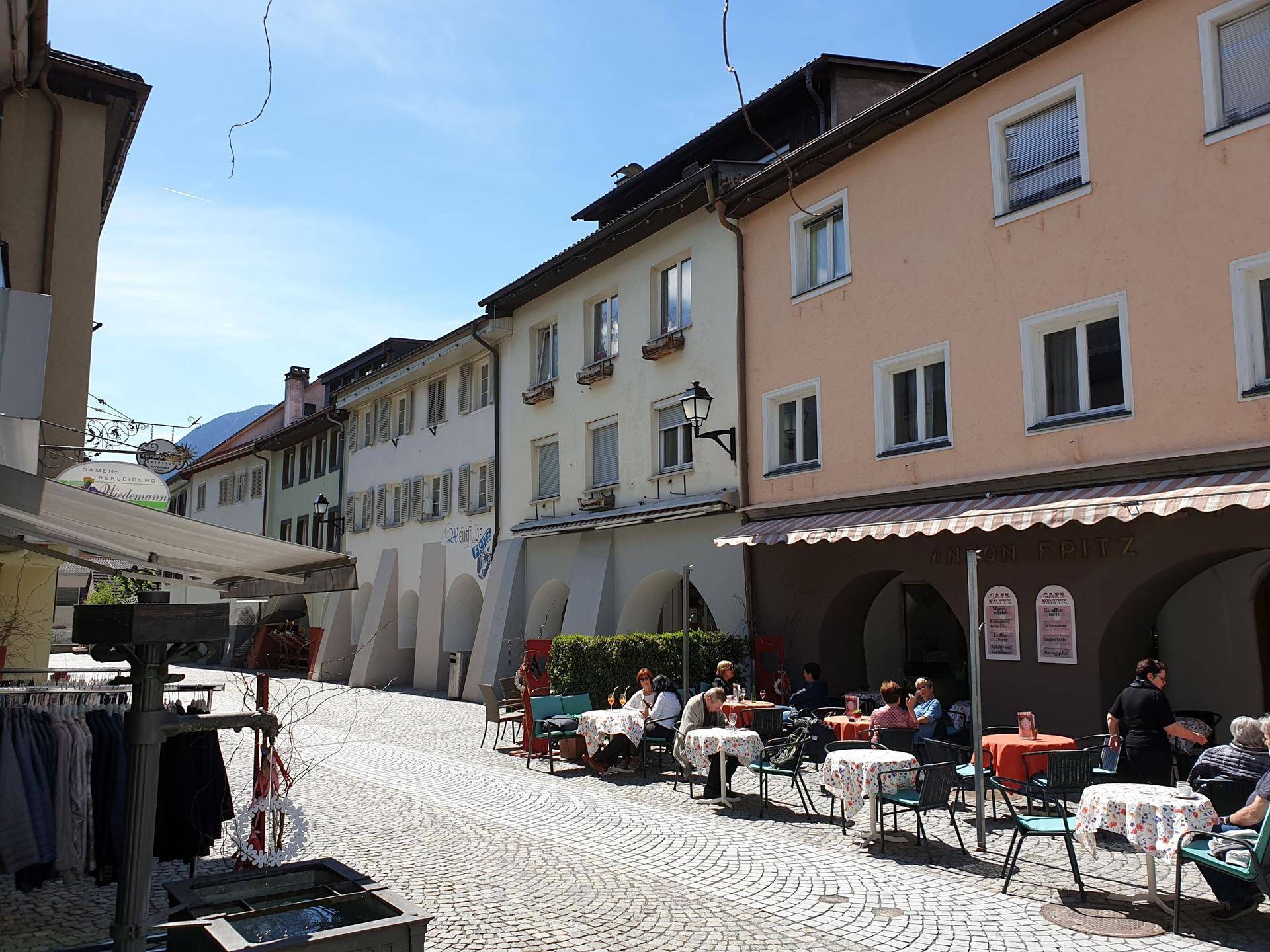 Belebtes Straßencafé mit Tischen und Stühlen im Freien, umgeben von traditionellen Gebäuden und Kopfsteinpflaster.