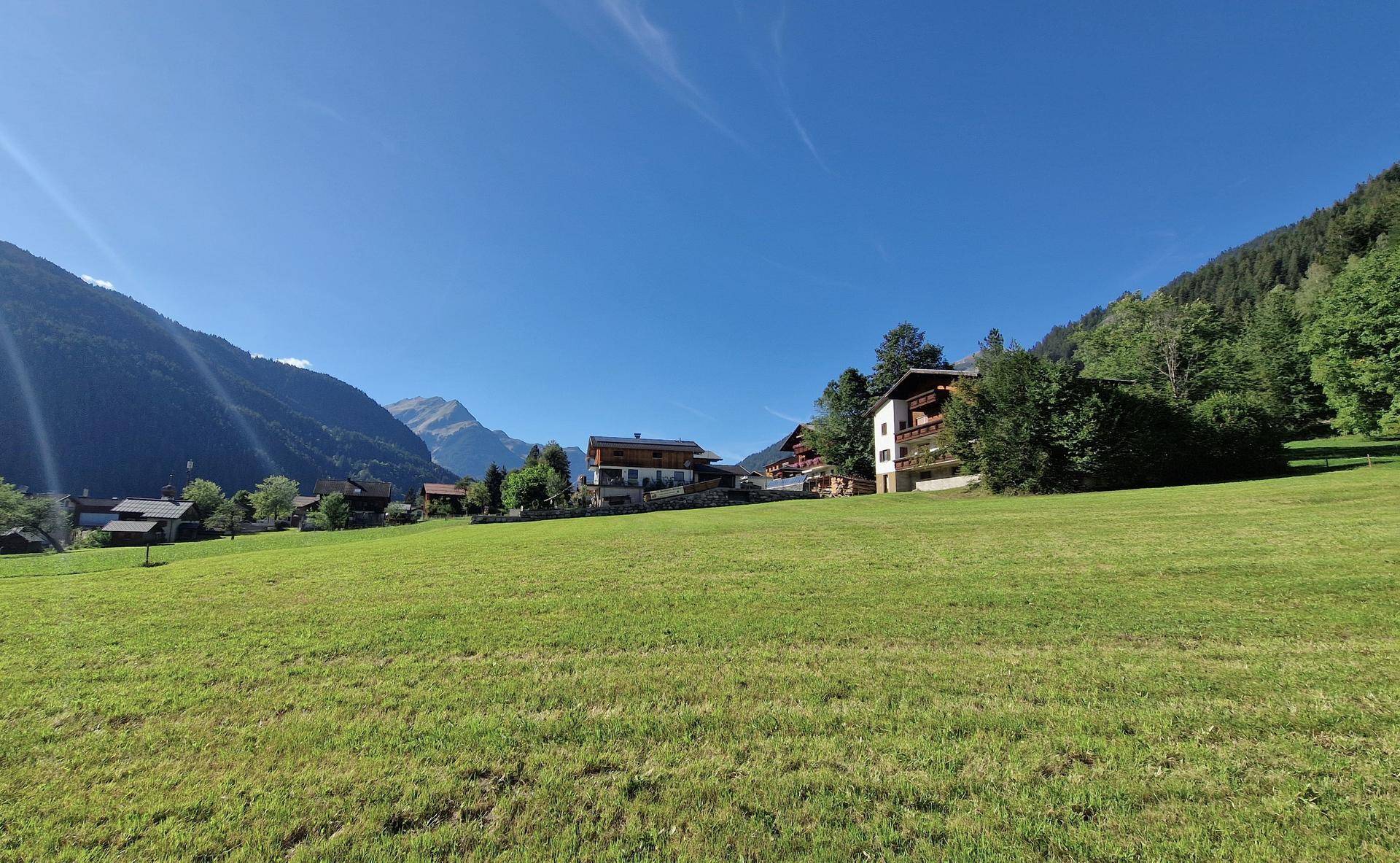 Sonnige grüne Wiese mit modernen Häusern im Hintergrund, umgeben von einer beeindruckenden Berglandschaft.