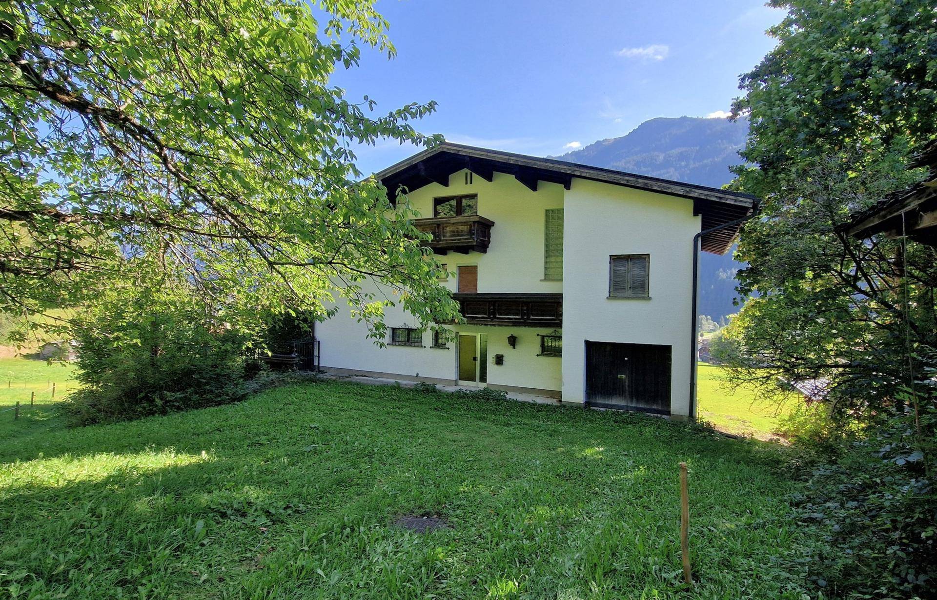 Vorderansicht des Hauses mit Garage und Balkonen, umgeben von üppiger Natur und Bergen.