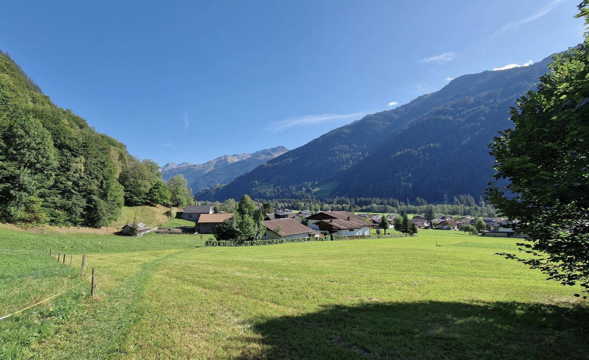 Weitläufige grüne Wiesen vor einem malerischen Dorf, eingebettet in ein Tal mit bewaldeten Bergen.