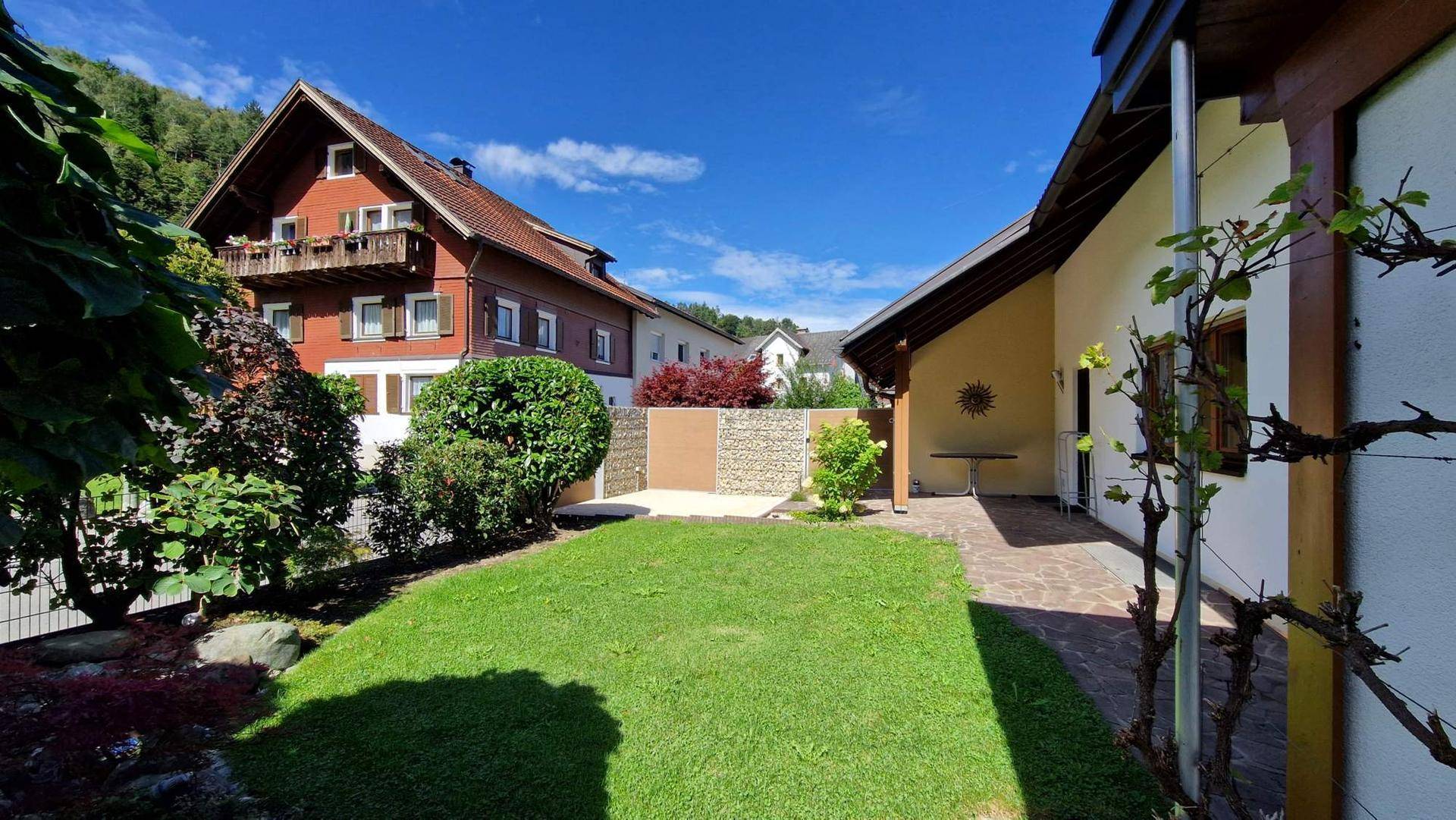 Gepflegter Garten mit Rasenfläche, Gabionenwand und Blick auf das Nachbargebäude.