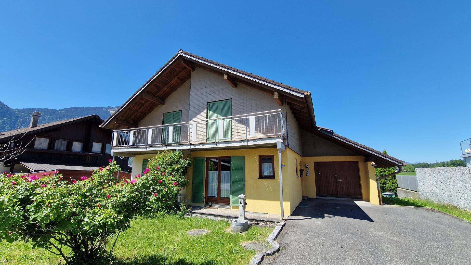 Ansicht des Hauses mit Balkon, Garage und Zufahrt, umgeben von Grünflächen und blühenden Büschen.