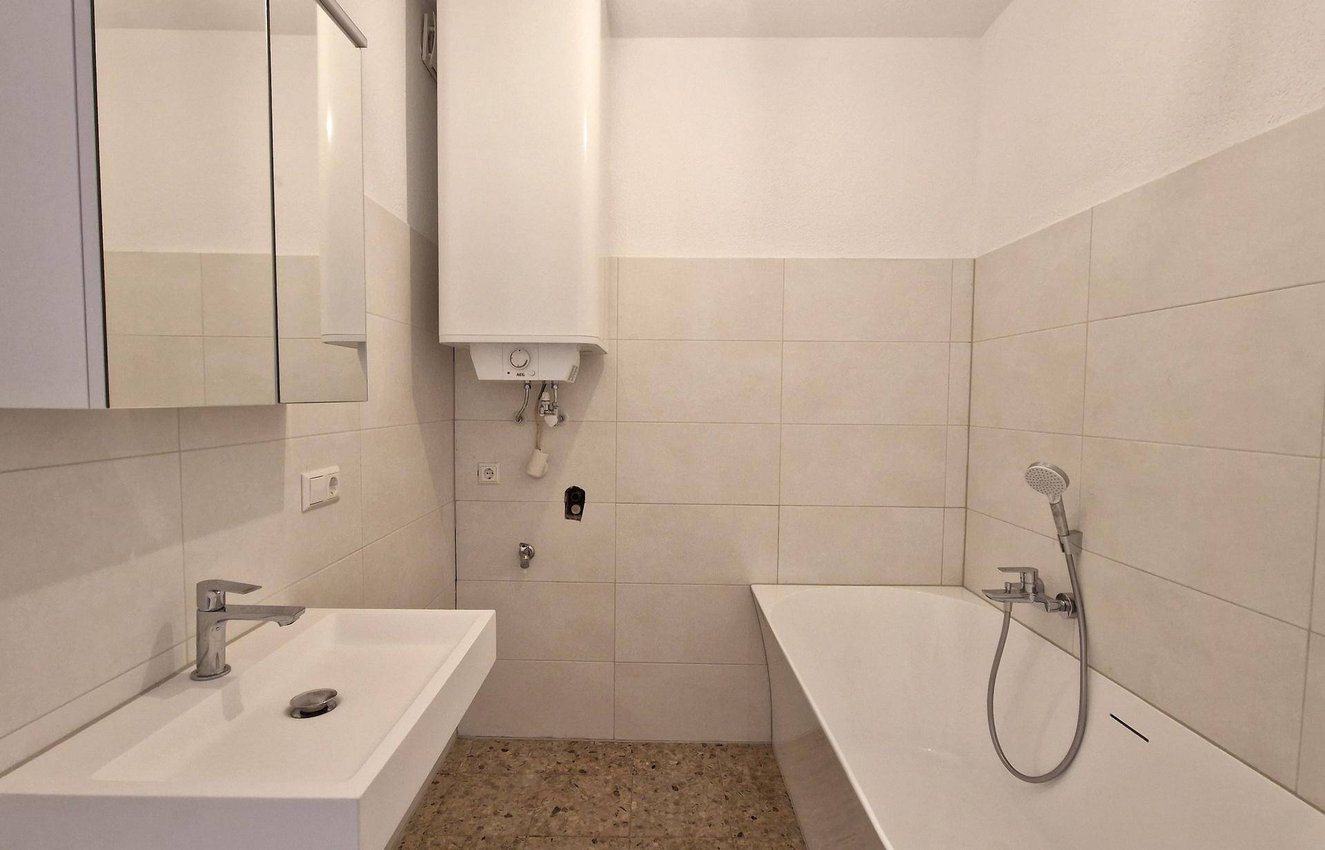 Helles Badezimmer mit Badewanne, Waschbecken und Boiler, teilweise gefliest und mit Terrazzoboden.