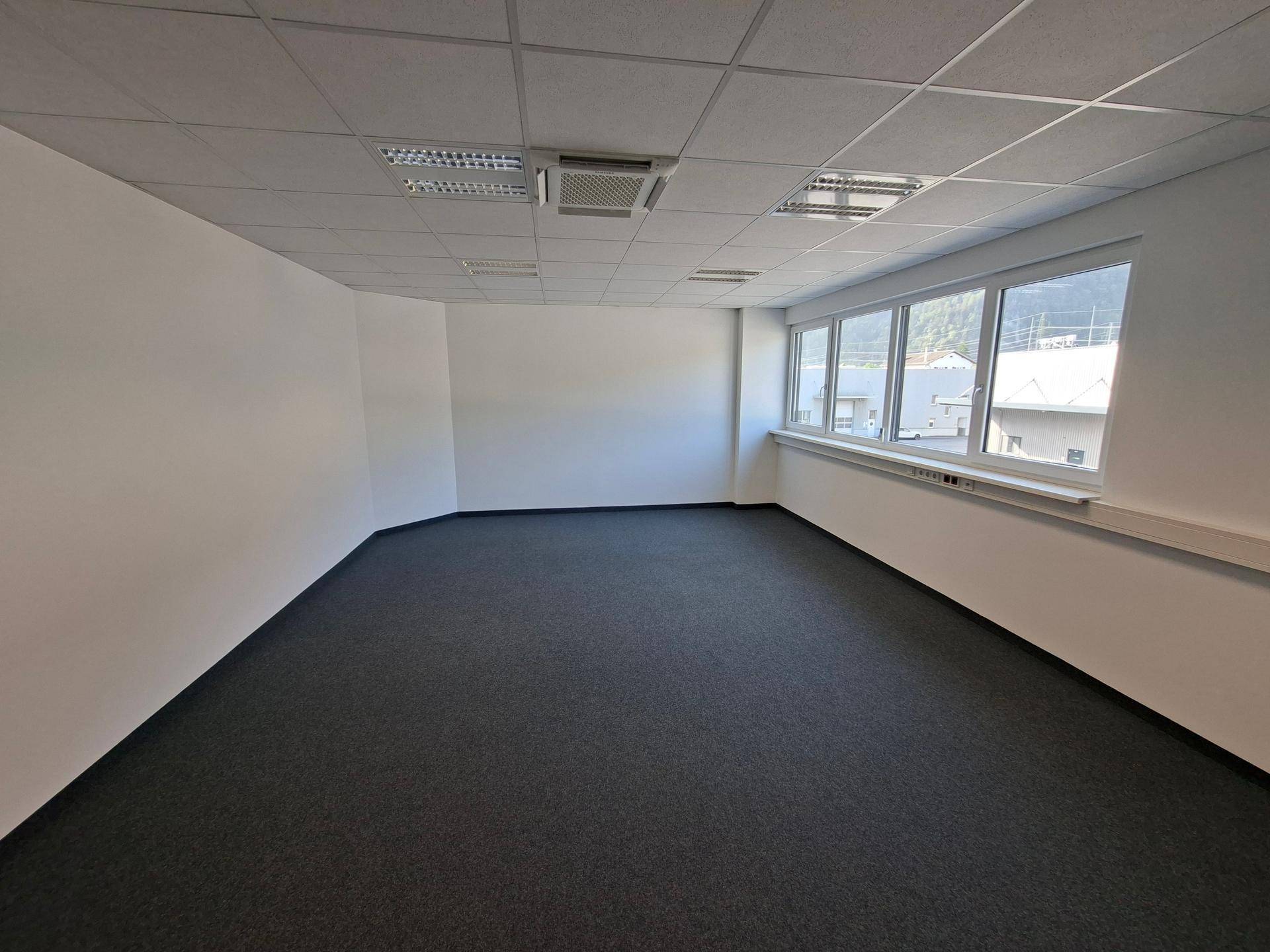 Geräumiges, leeres Büro mit großen Fenstern, grauem Teppichboden und abgehängter Decke.