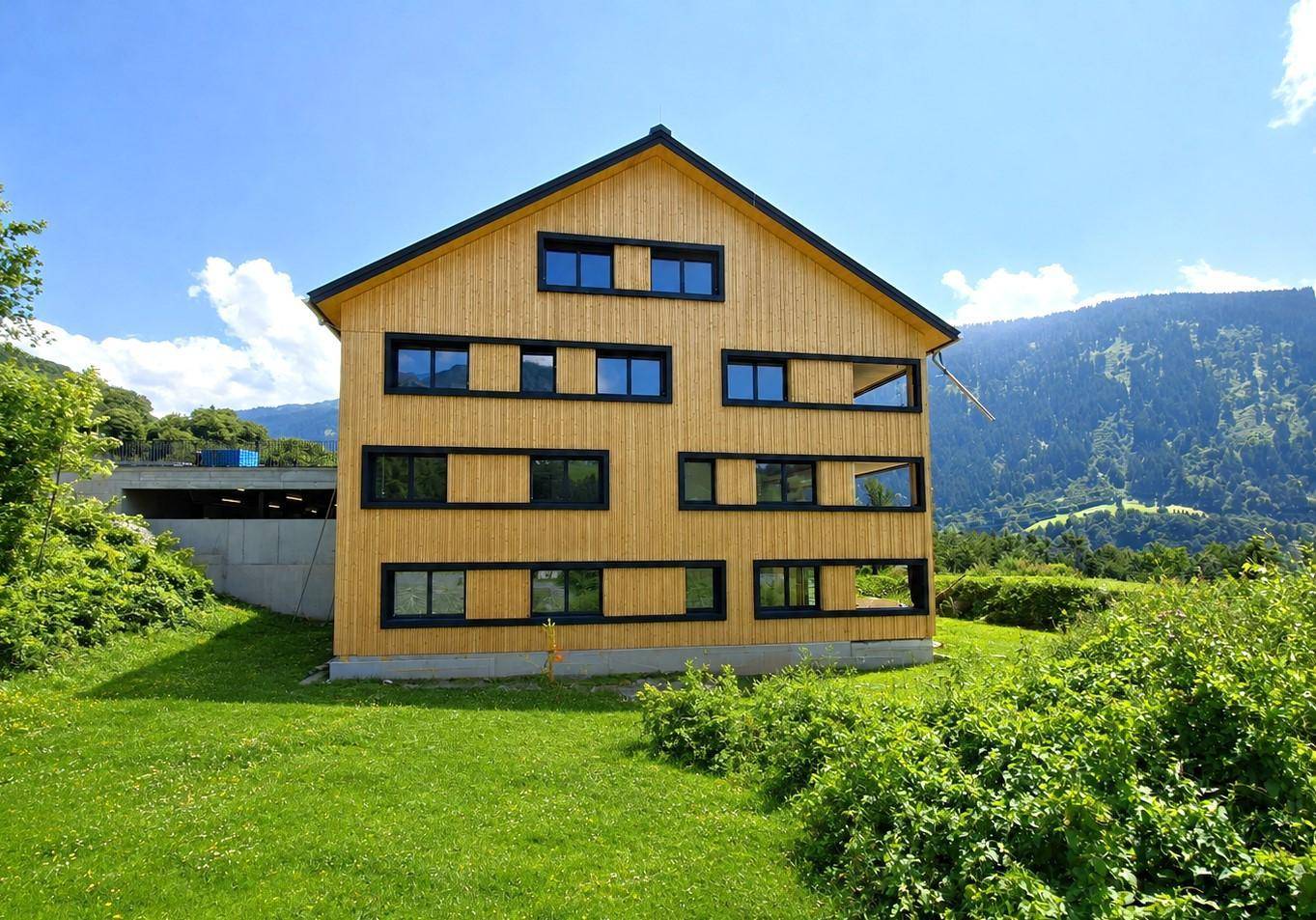 Seitenansicht des modernen Holzgebäudes mit Giebeldach und großen Fenstern, umgeben von Natur.