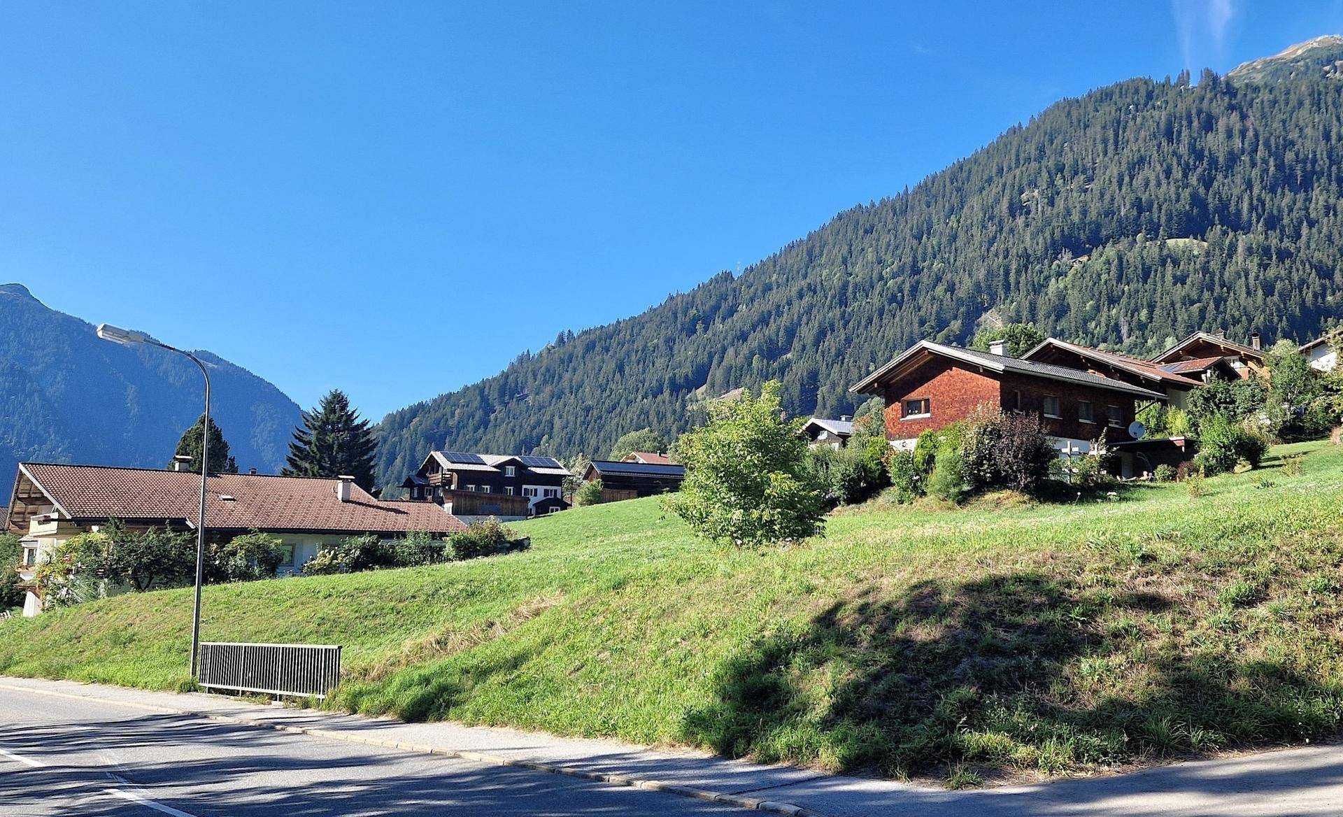 Häuser am Hang mit Blick auf die Berge, umgeben von grünen Wiesen und einer Straße im Vordergrund.