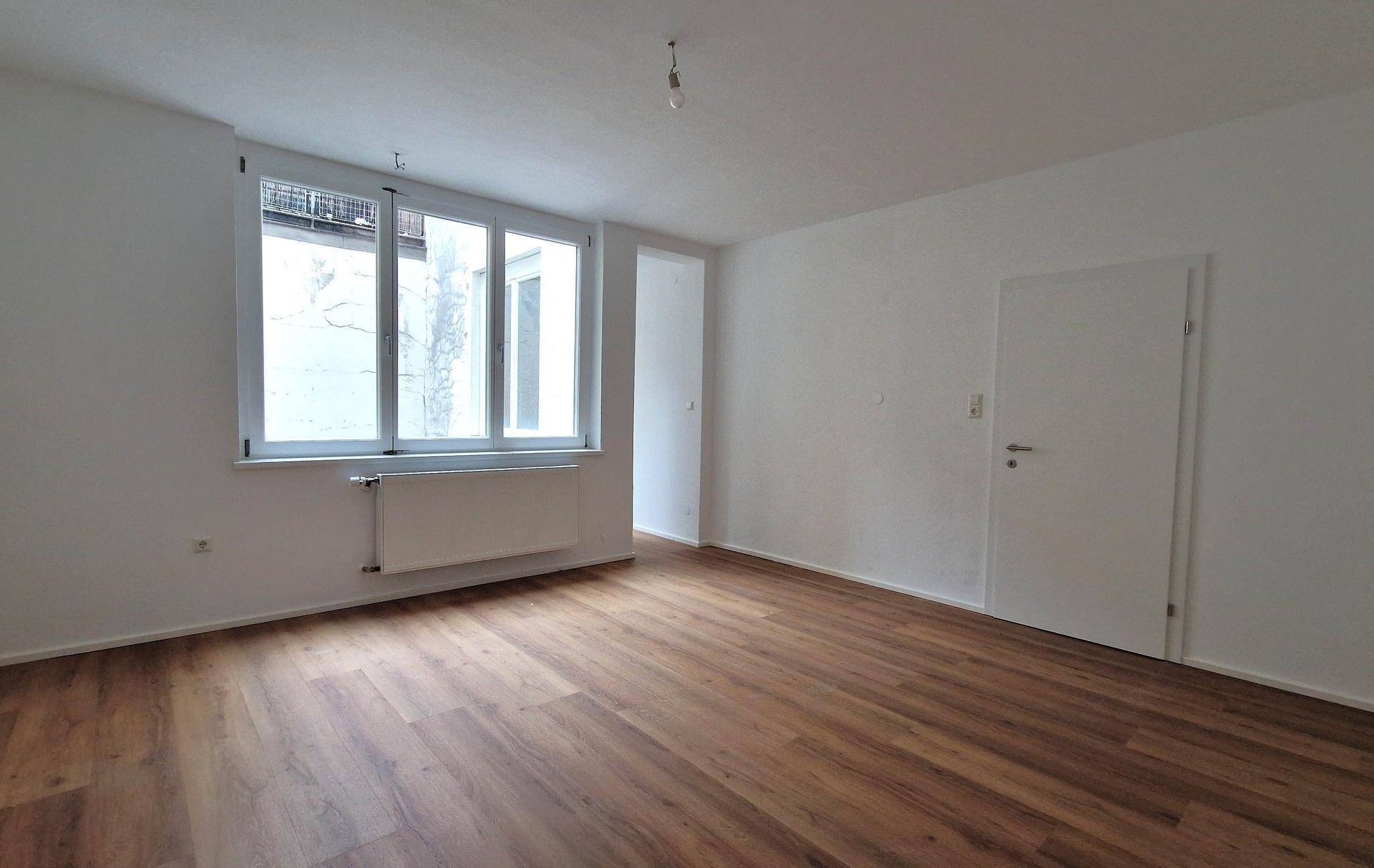 Geräumiger Wohnraum mit Laminatboden, weißen Wänden und einem großen Fenster mit Blick auf einen Innenhof.