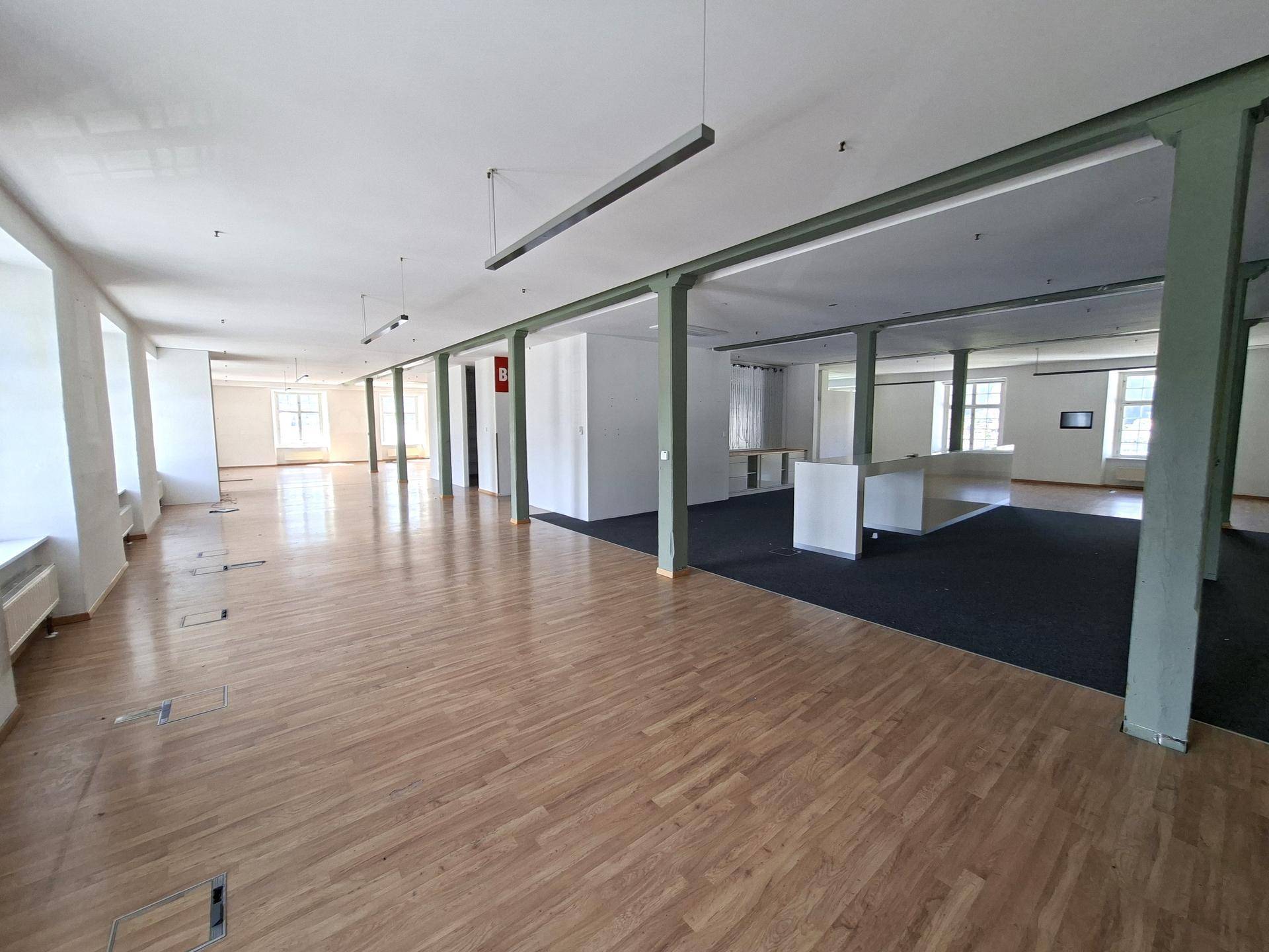 Weitläufiger Bürobereich mit Holzboden, Teppichbereichen und grünen Stützpfeilern.