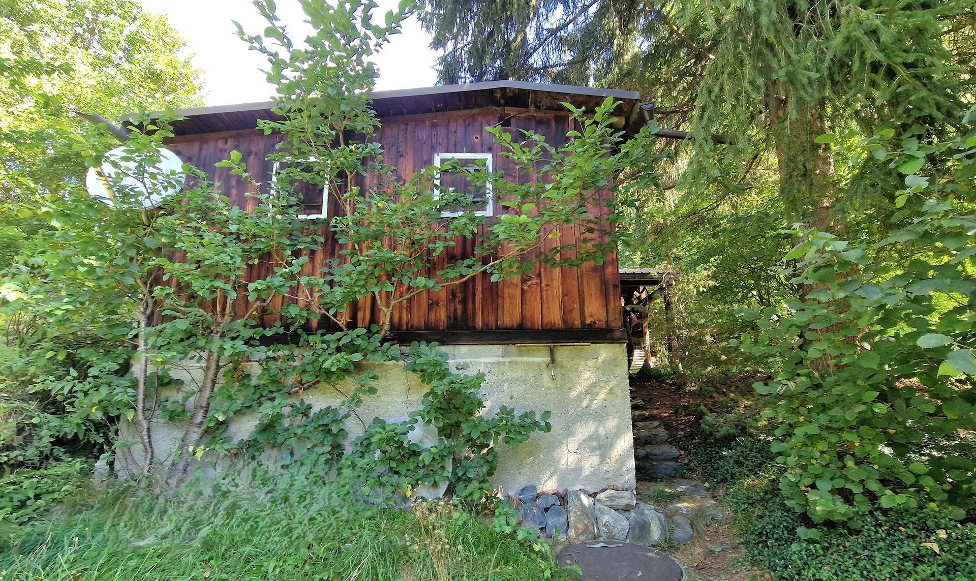 Rustikales Holzhaus mit einem Steinfundament, eingebettet in üppiges Grün und hohe Bäume.