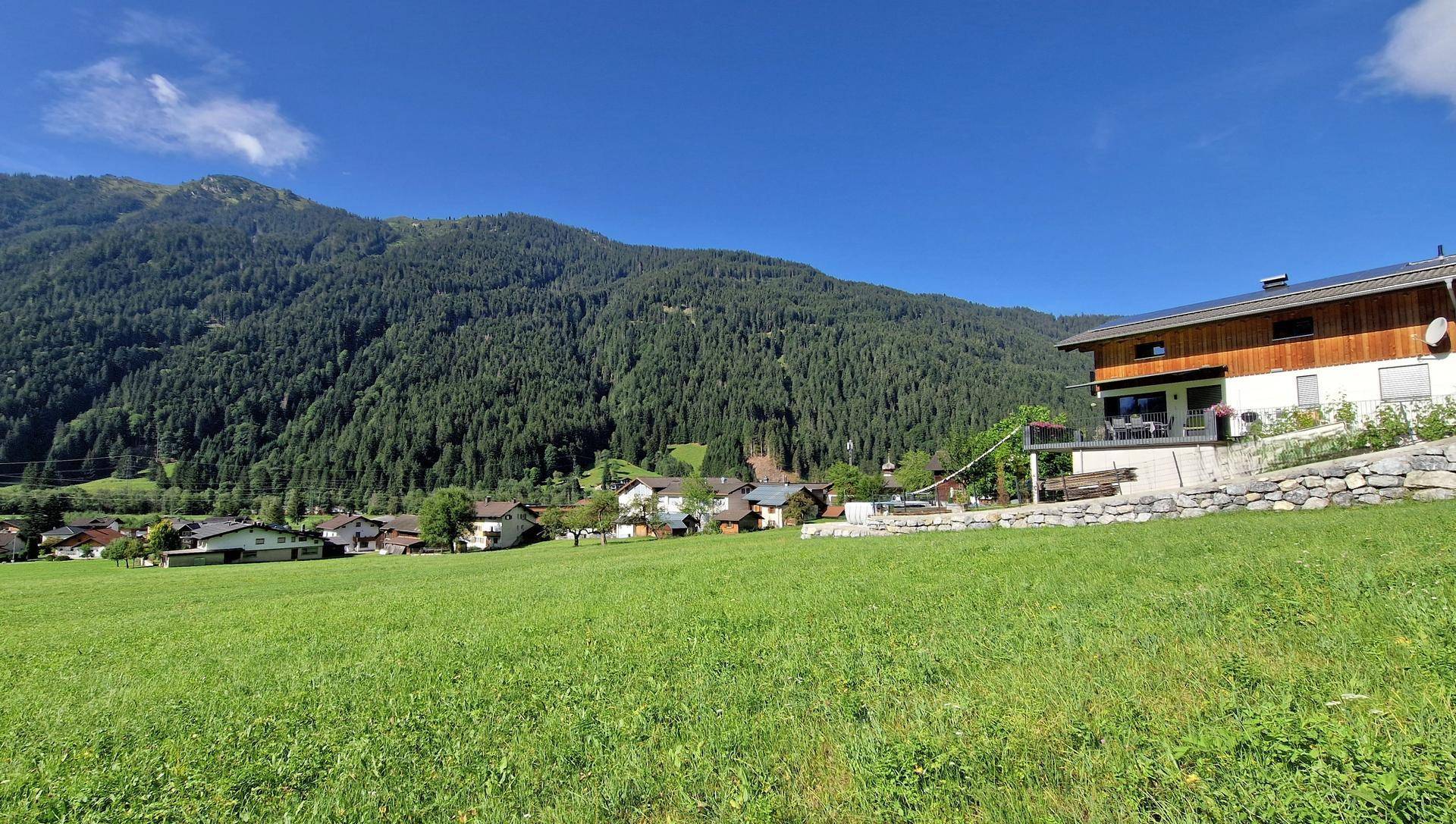 Modernes Haus mit Balkon und Blick über grüne Wiesen auf ein Dorf und die umliegenden Berge.