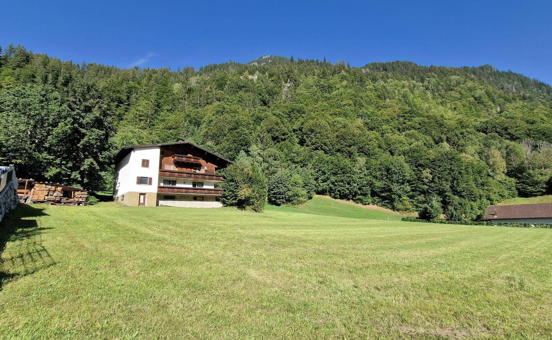 Weißes Haus mit Holzbalkonen auf einer weitläufigen grünen Wiese vor einem dichten Wald und Bergen.