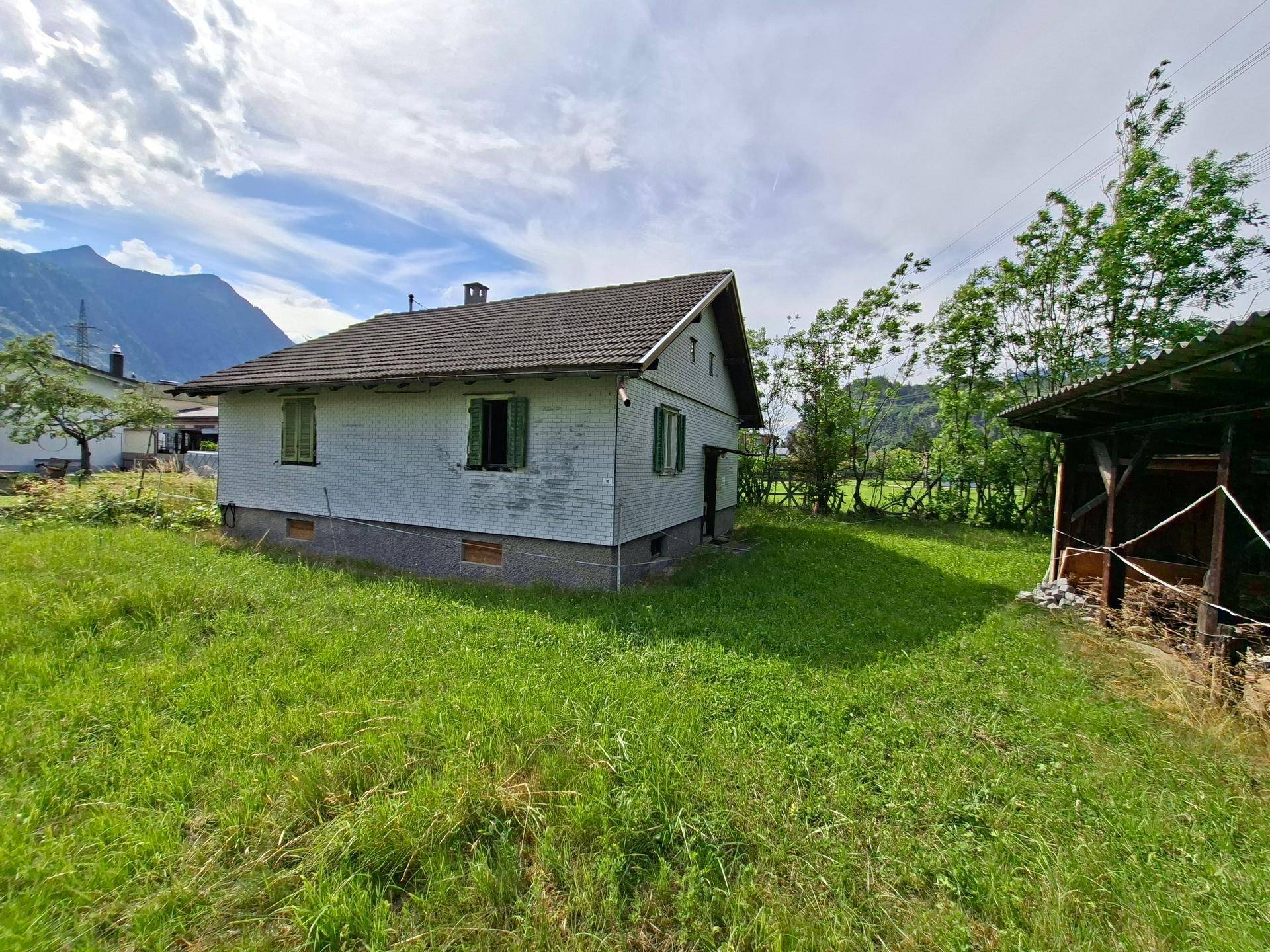 Seitenansicht eines älteren Hauses mit Satteldach und einem kleinen Schuppen, umgeben von Grünflächen.