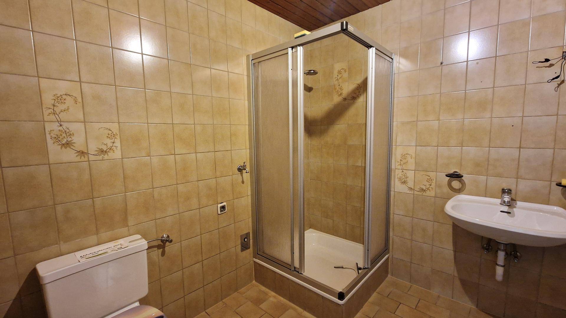 Badezimmer mit beigefarbenen Fliesen, Duschkabine und einem kleinen Waschbecken.
