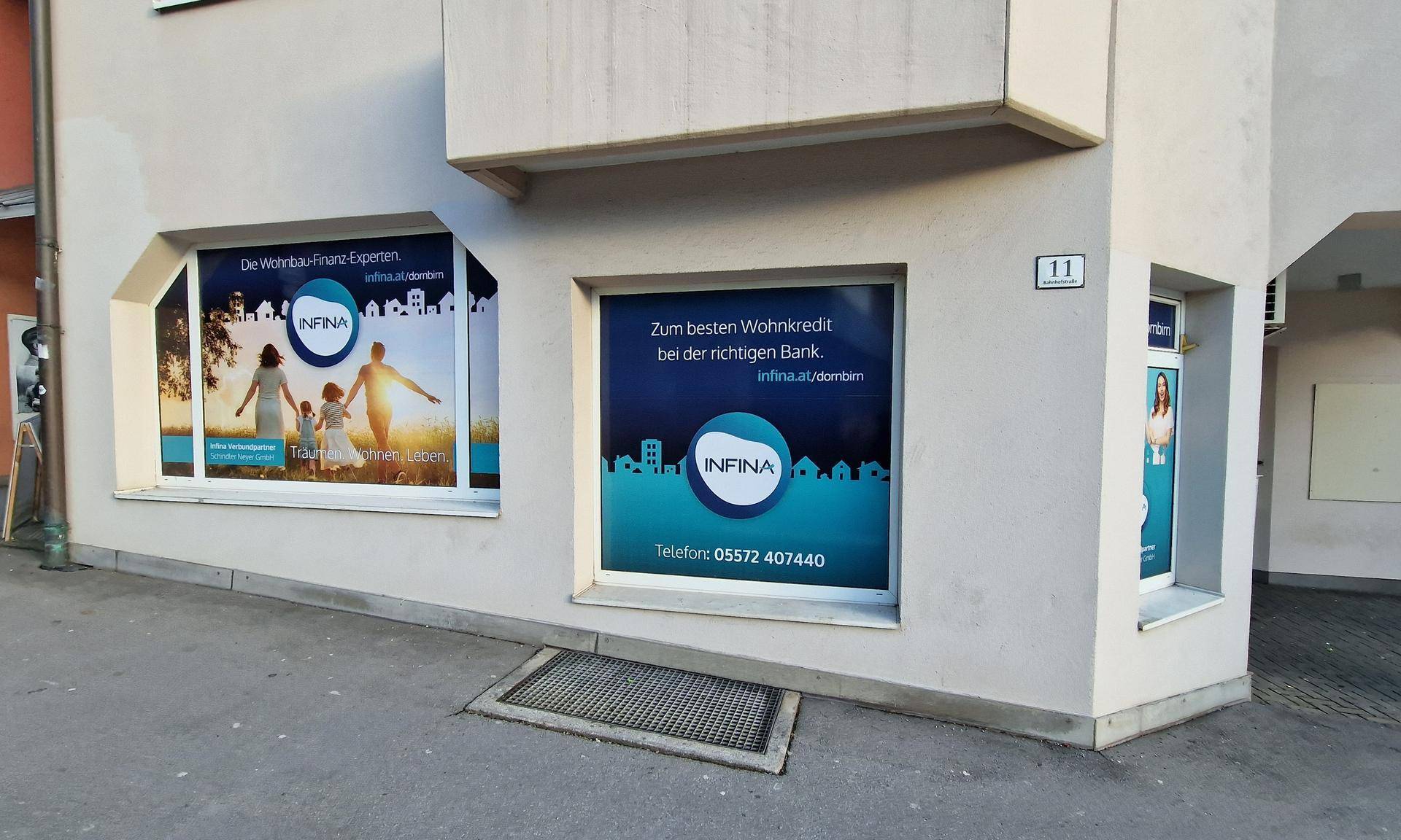 Detaillierte Ansicht der Gebäudefassade mit großen Schaufenstern, die Werbeplakate zeigen, und einem Gitterrost am Boden.