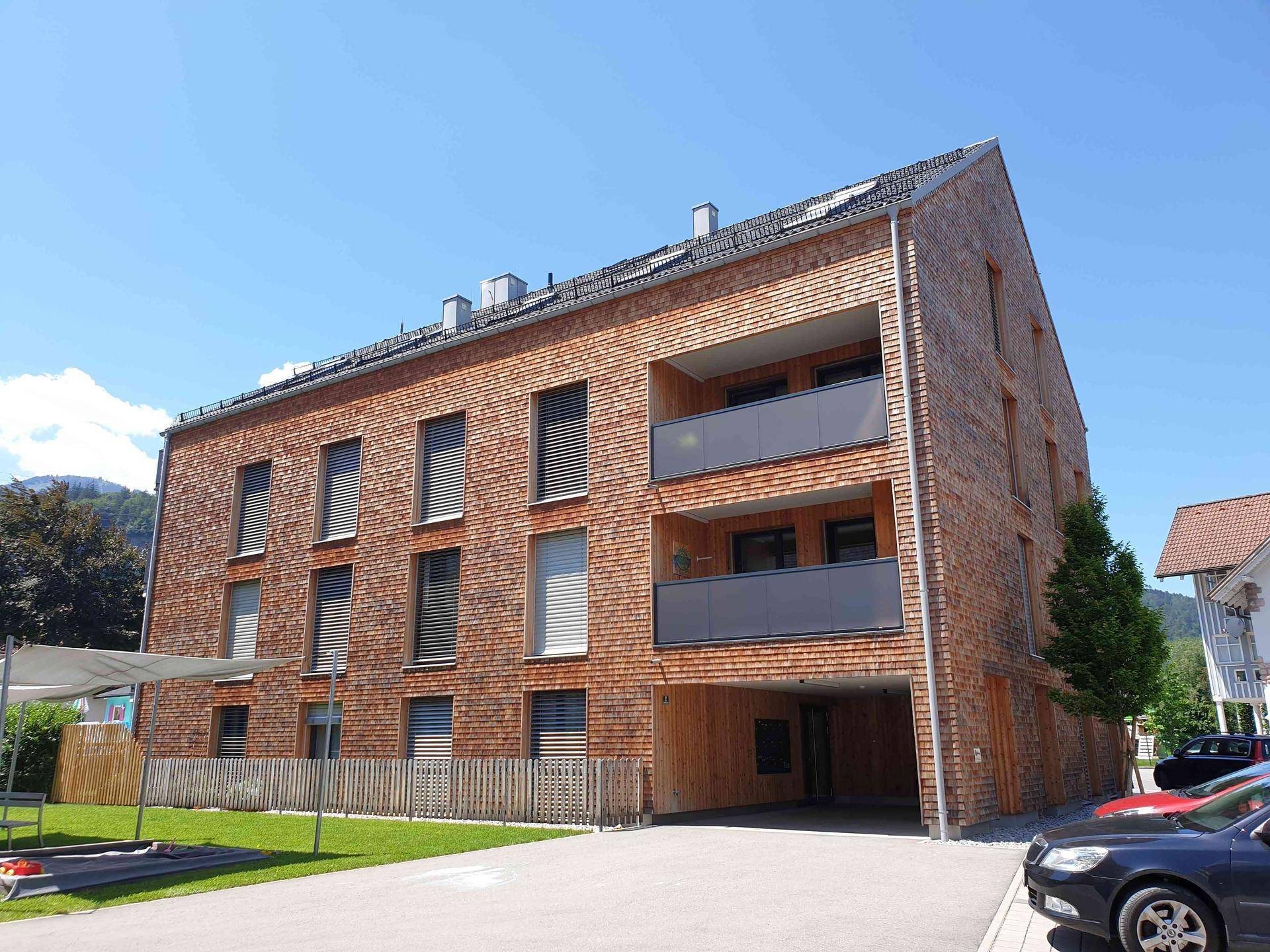 Neubau mit Holzfassade, Balkonen und überdachtem Parkplatz, umgeben von Grünflächen.