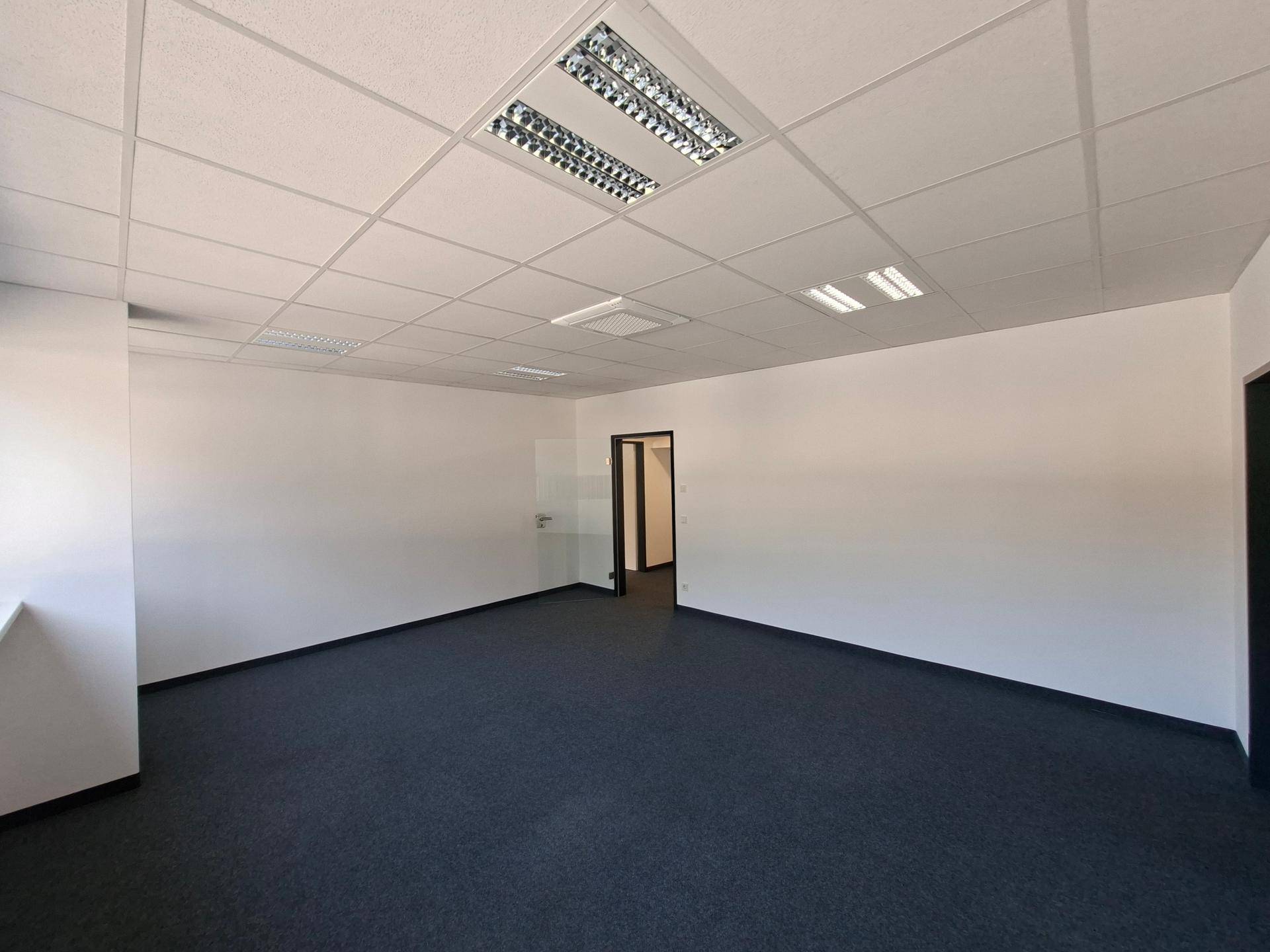 Leerer Büroraum mit grauem Teppichboden, abgehängter Decke und Zugang zu weiteren Räumen.