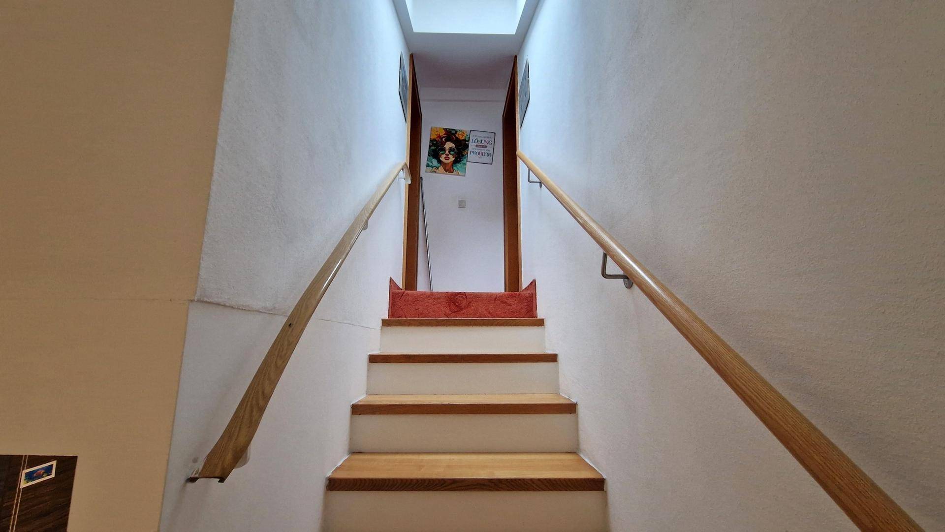 Holztreppe mit Geländer führt in die obere Etage, gut beleuchtet durch Oberlicht.