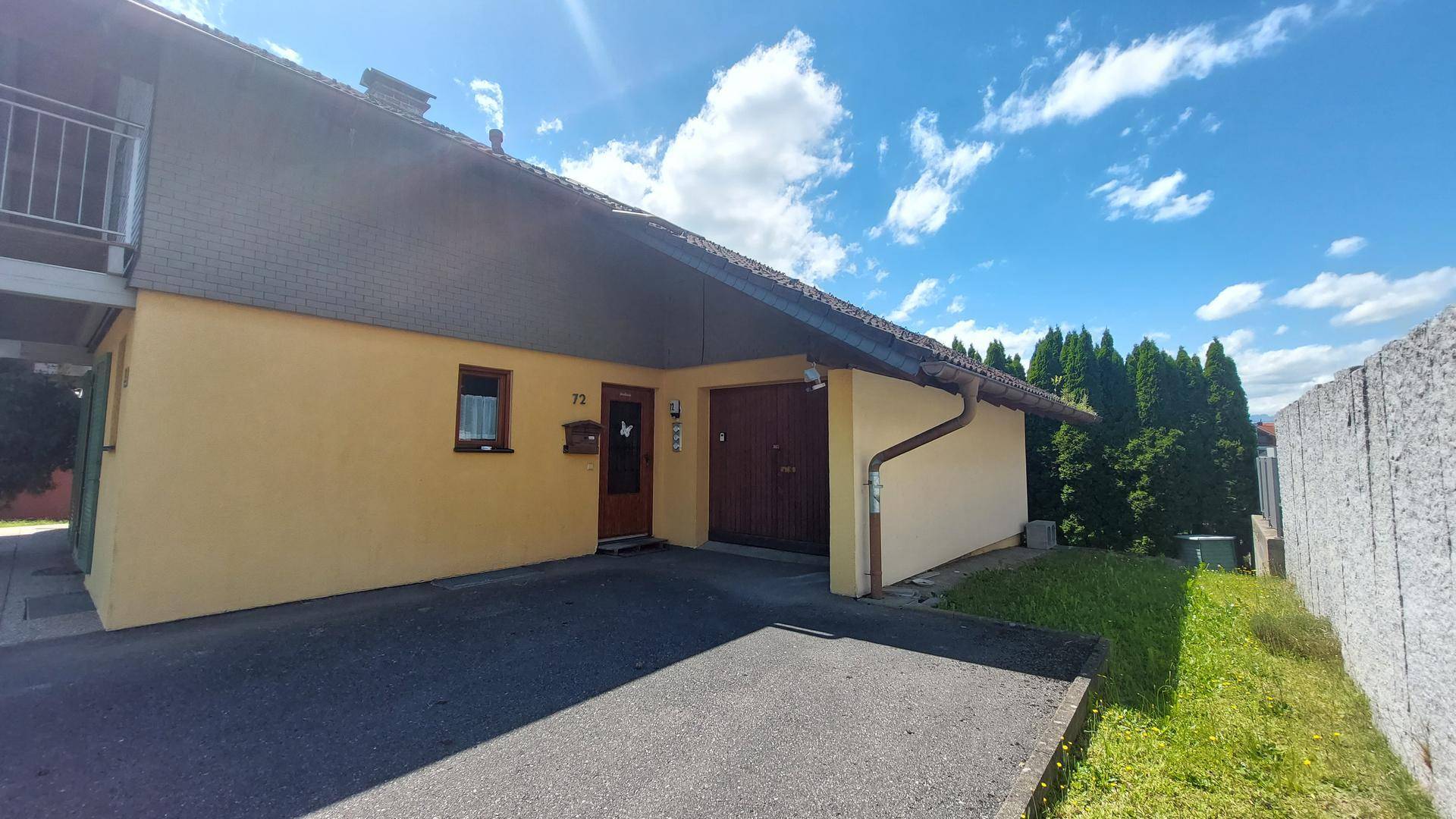 Seitenansicht des Hauses mit Garage, Eingangsbereich und asphaltierter Zufahrt unter blauem Himmel.