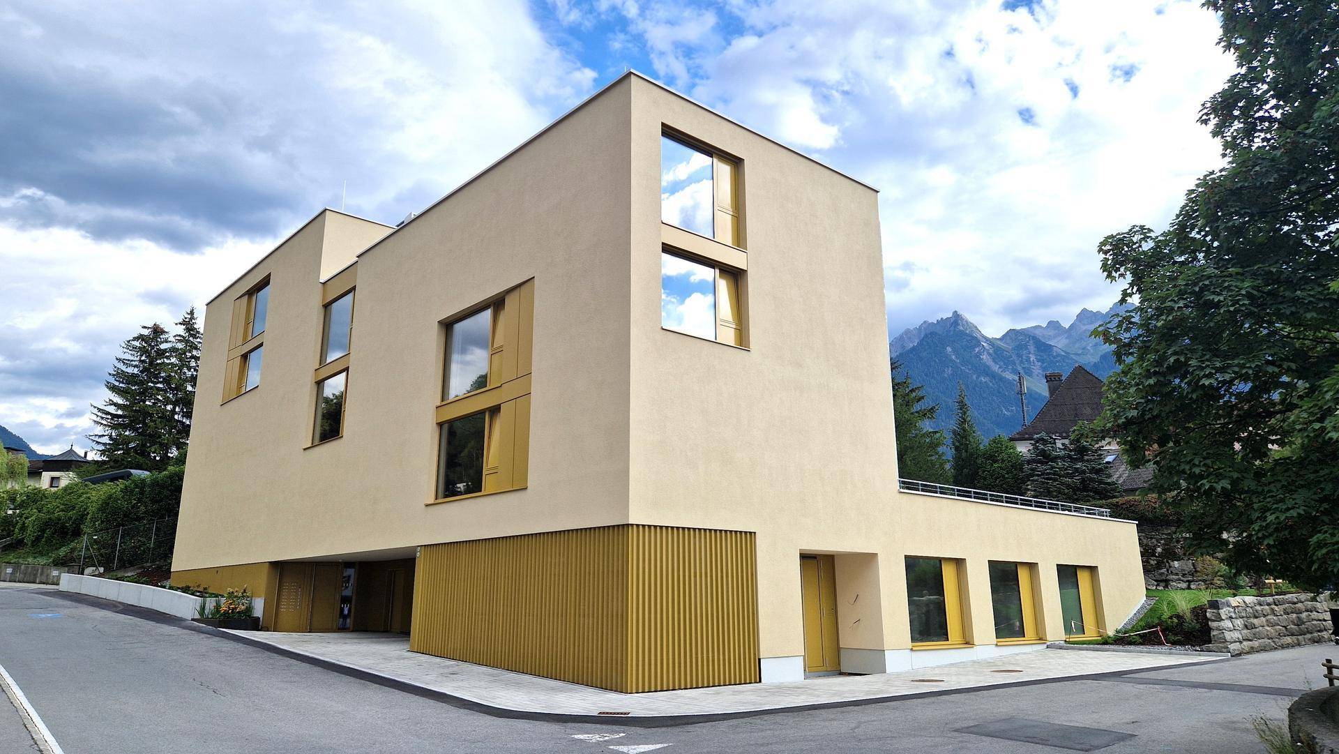 Ansicht des modernen Gebäudes mit beiger Fassade und Holzverkleidung im unteren Bereich, umgeben von Bergen.