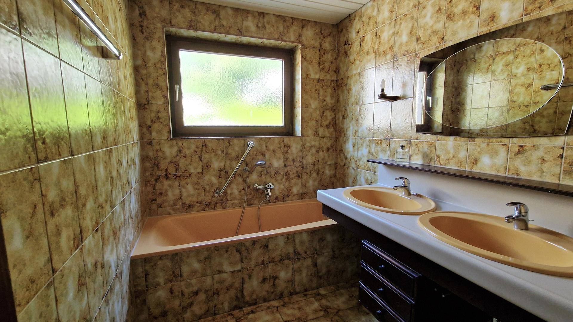 Badezimmer im 80er-Jahre-Stil mit beigefarbenen Fliesen, Badewanne und Doppelwaschbecken.
