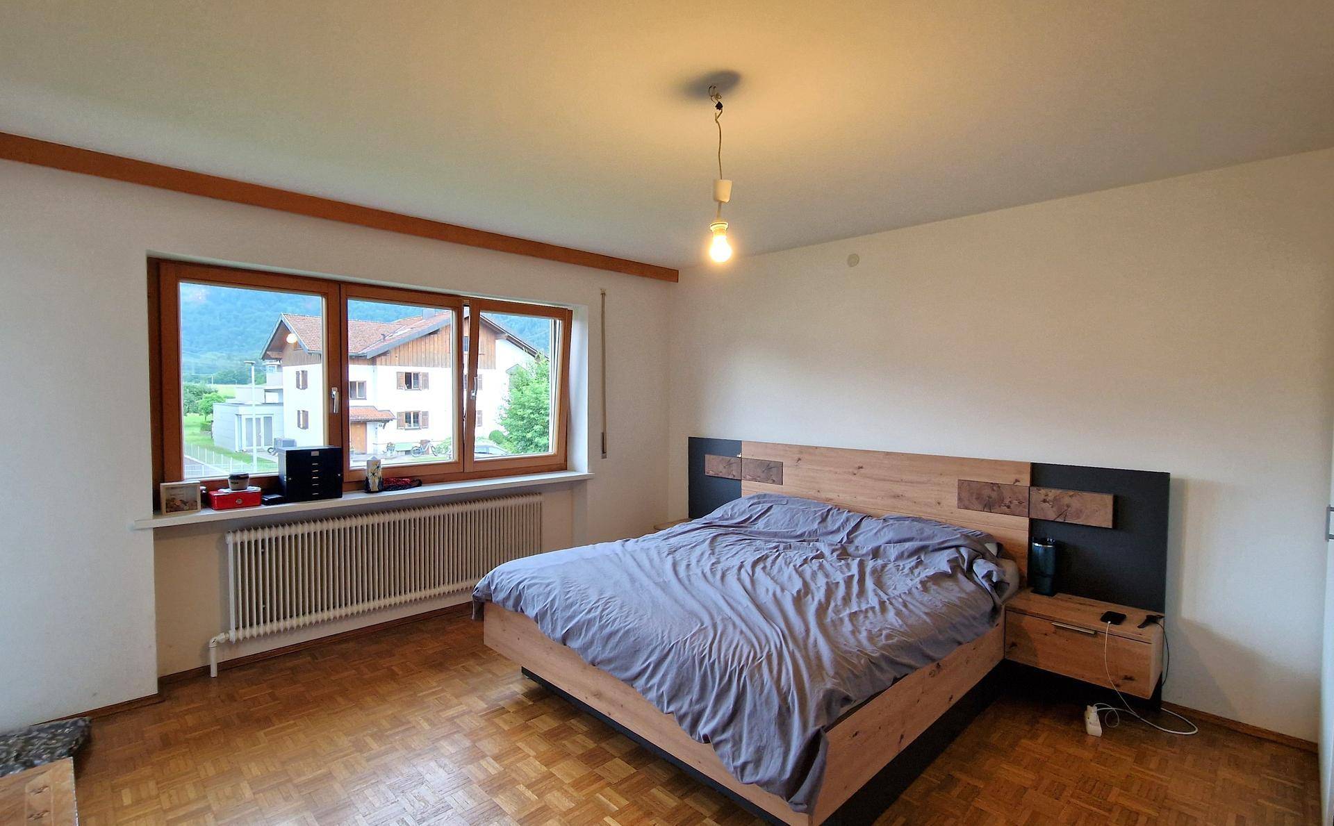 Helles Schlafzimmer mit Holzbett, Parkettboden und großem Fenster mit Blick ins Grüne.