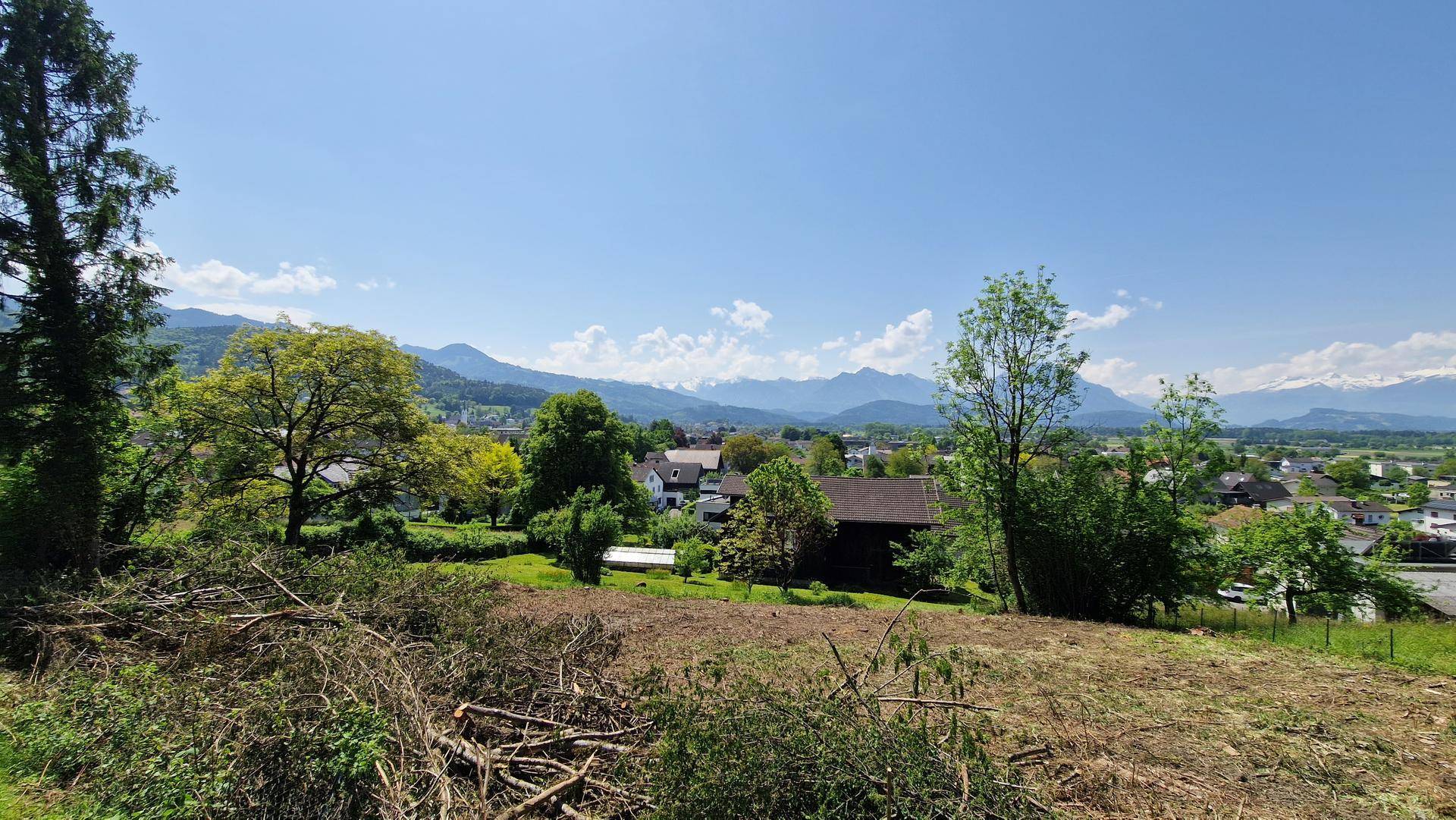 Sonniger Ausblick über ein Grundstück mit Blick auf eine Siedlung und die Alpenkette.