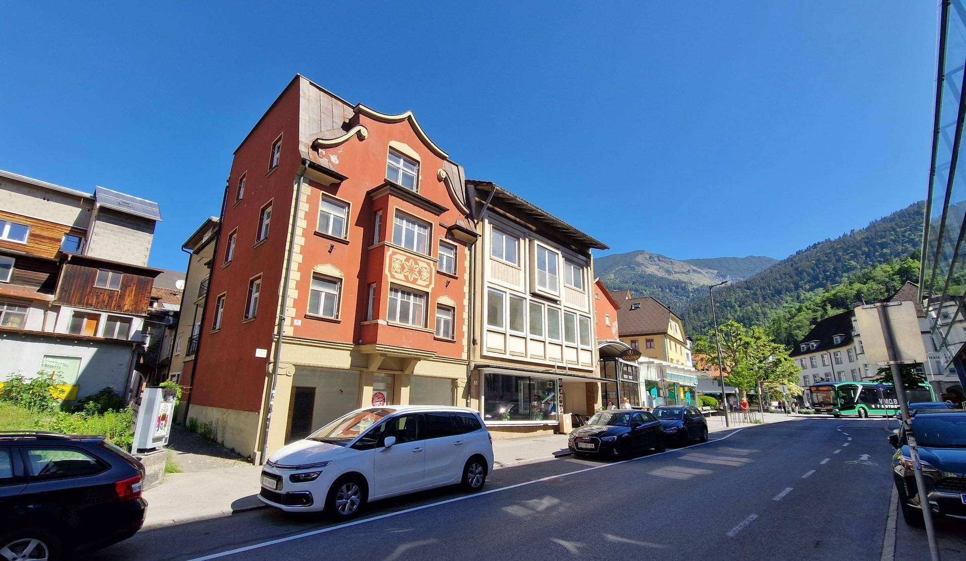 Gebäudeansicht an einer Straße mit parkenden Autos und Bergen im Hintergrund unter blauem Himmel.