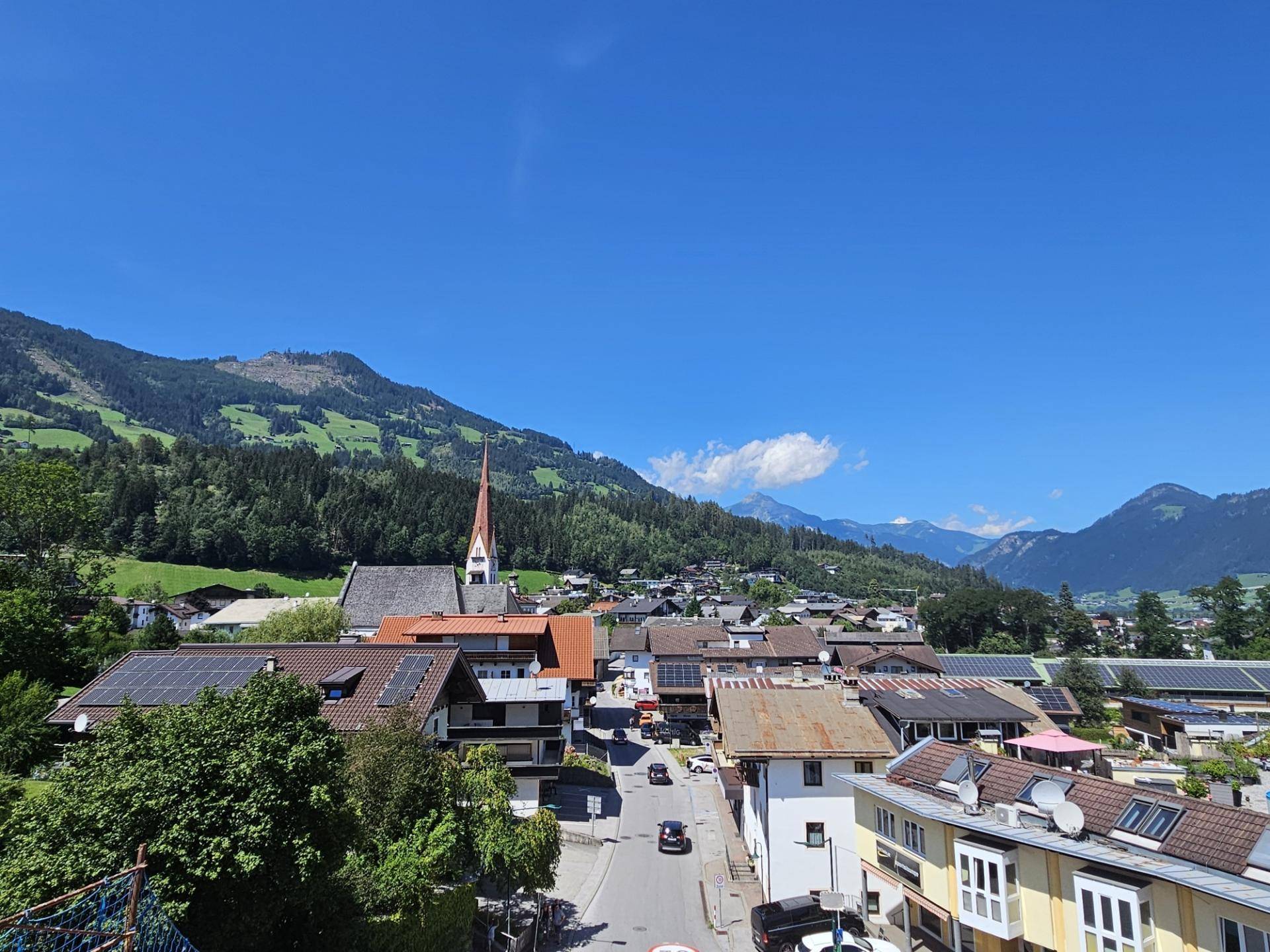 Panoramablick über ein malerisches Dorf mit Kirche, umgeben von grünen Bergen und einem klaren blauen Himmel.