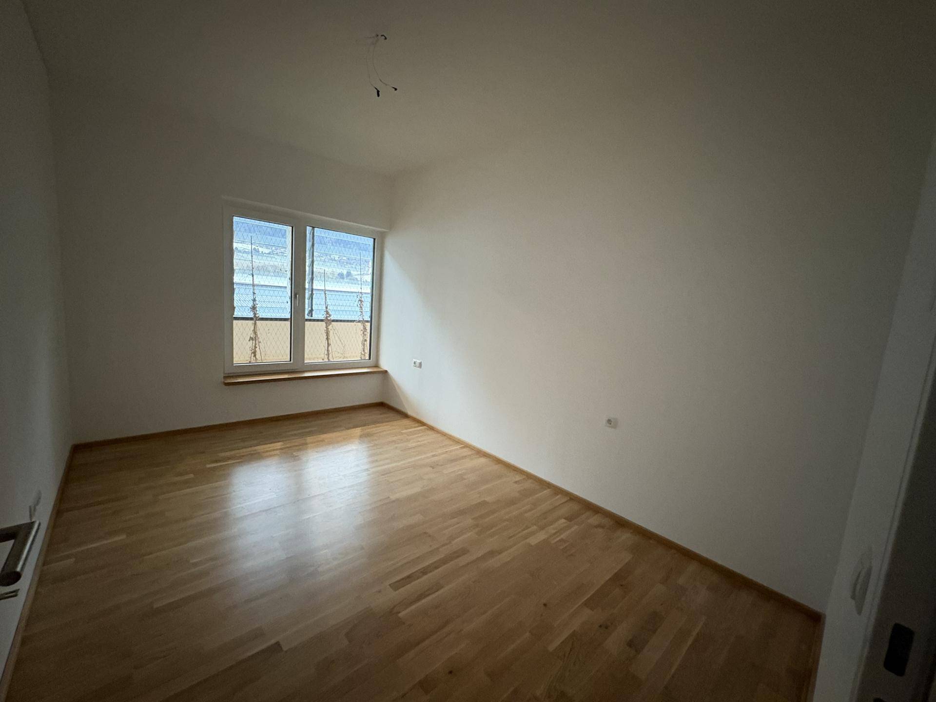 Leeres Zimmer mit Parkettboden und großem Fenster, ideal als Schlaf- oder Arbeitszimmer.