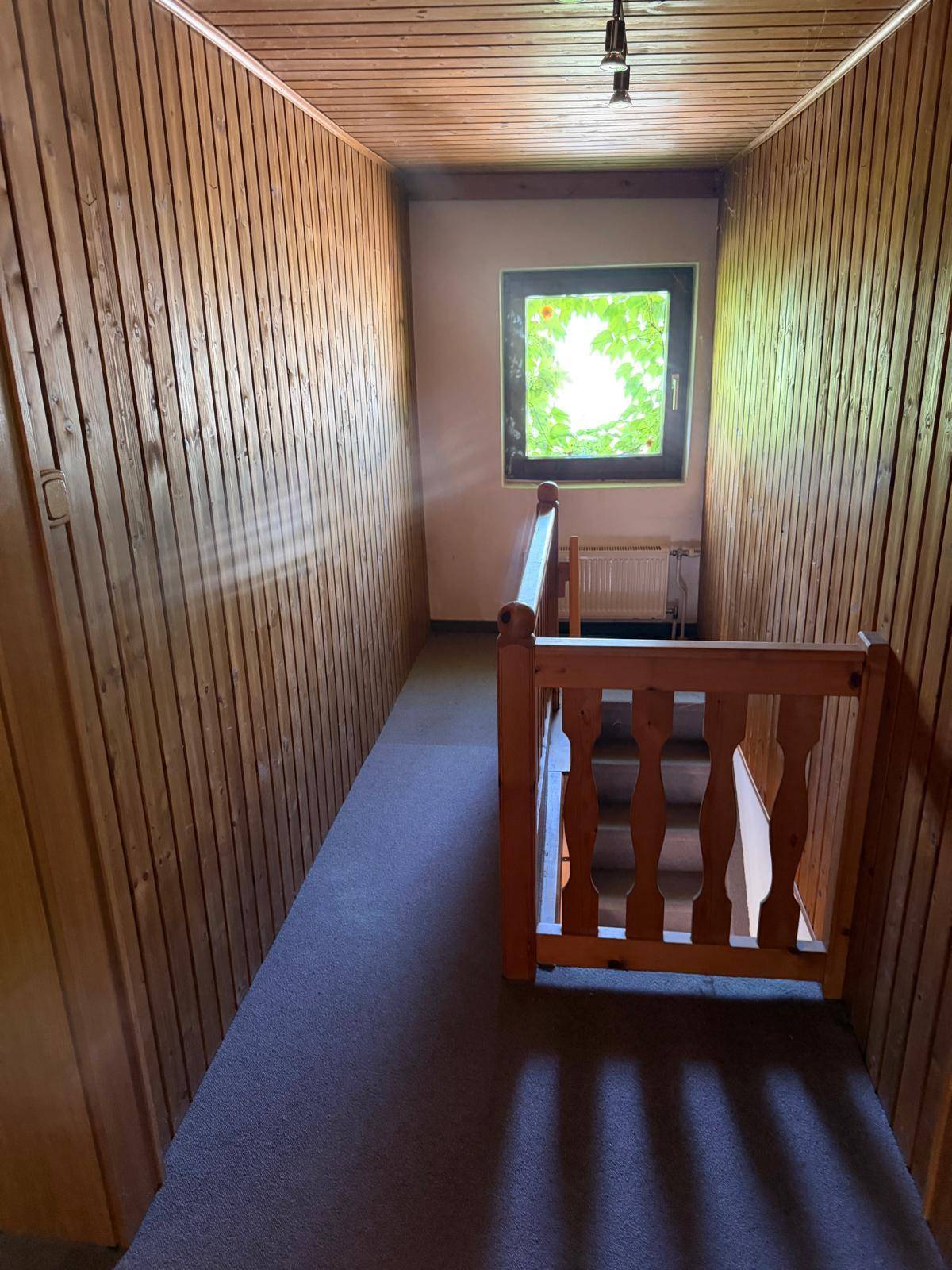 Flur mit Holzvertäfelung, Teppichboden und Treppengeländer, beleuchtet durch ein Fenster.