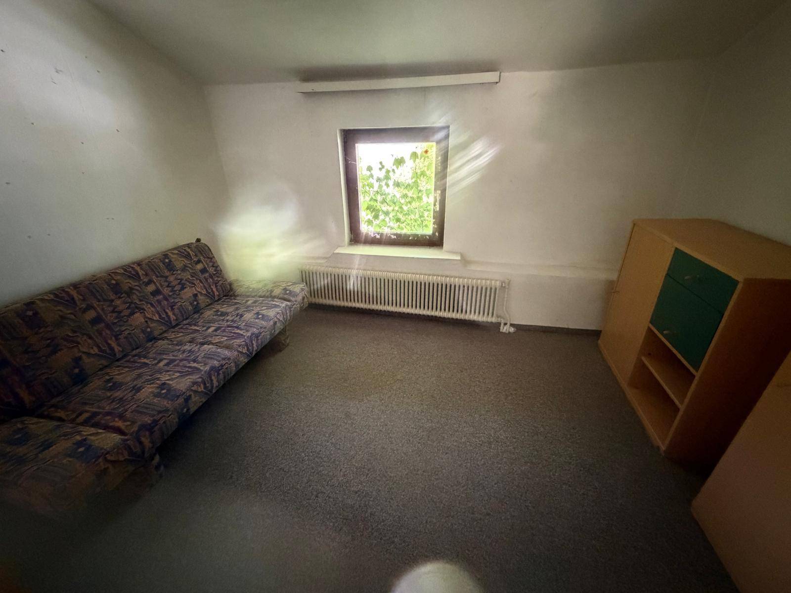 Einfacher Raum mit Teppichboden, Sofa und einem Fenster, das Tageslicht hereinlässt.