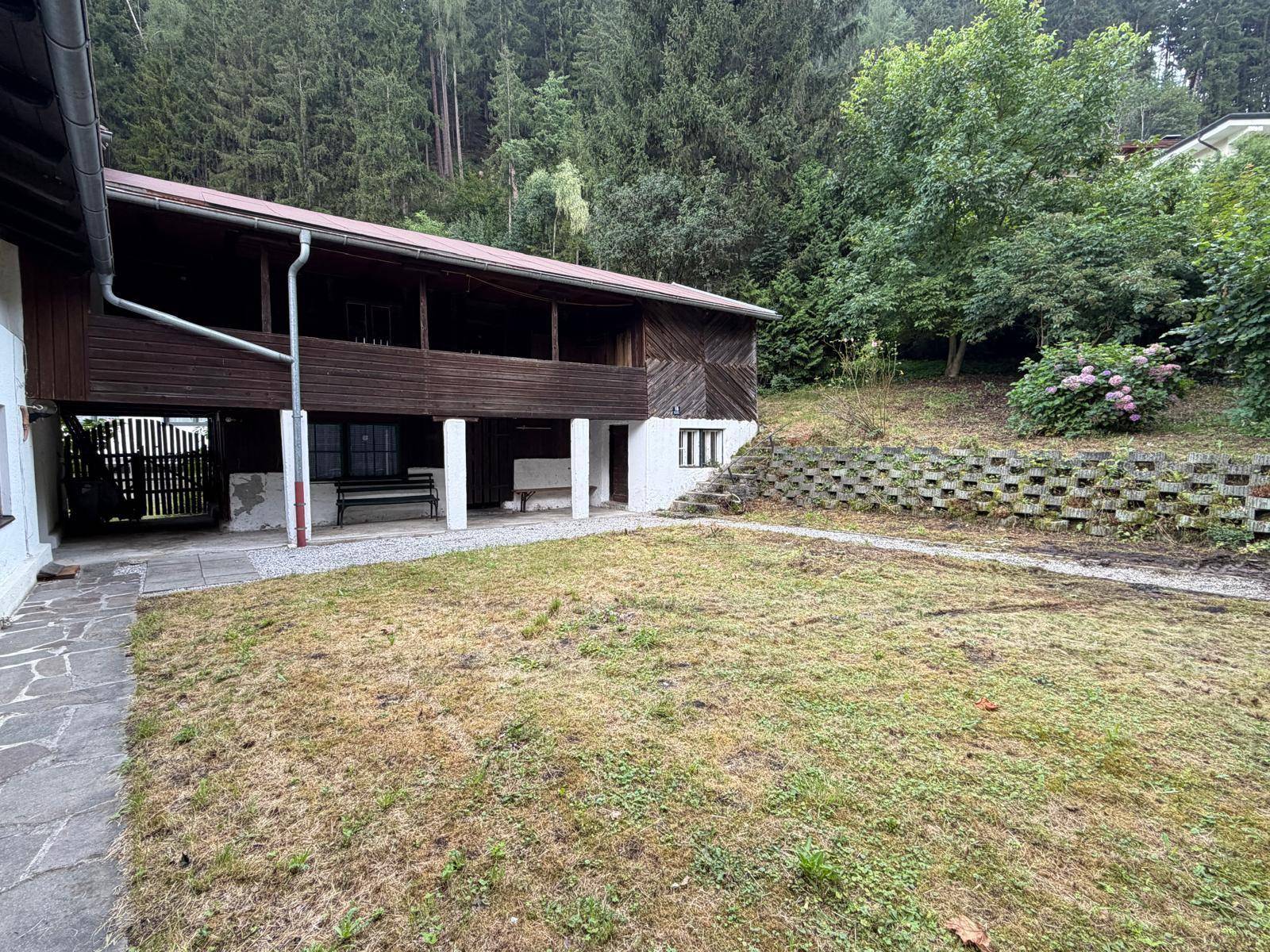 Rustikales Holzhaus mit überdachter Loggia und Gartenbereich vor bewaldetem Hang.