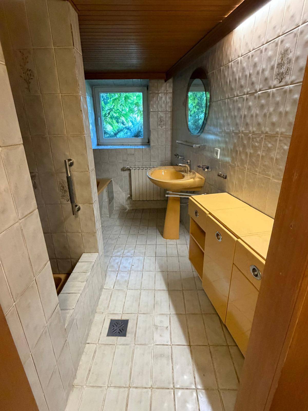 Badezimmer mit beigen Fliesen, gelbem Waschbecken, Badewanne und Fenster.