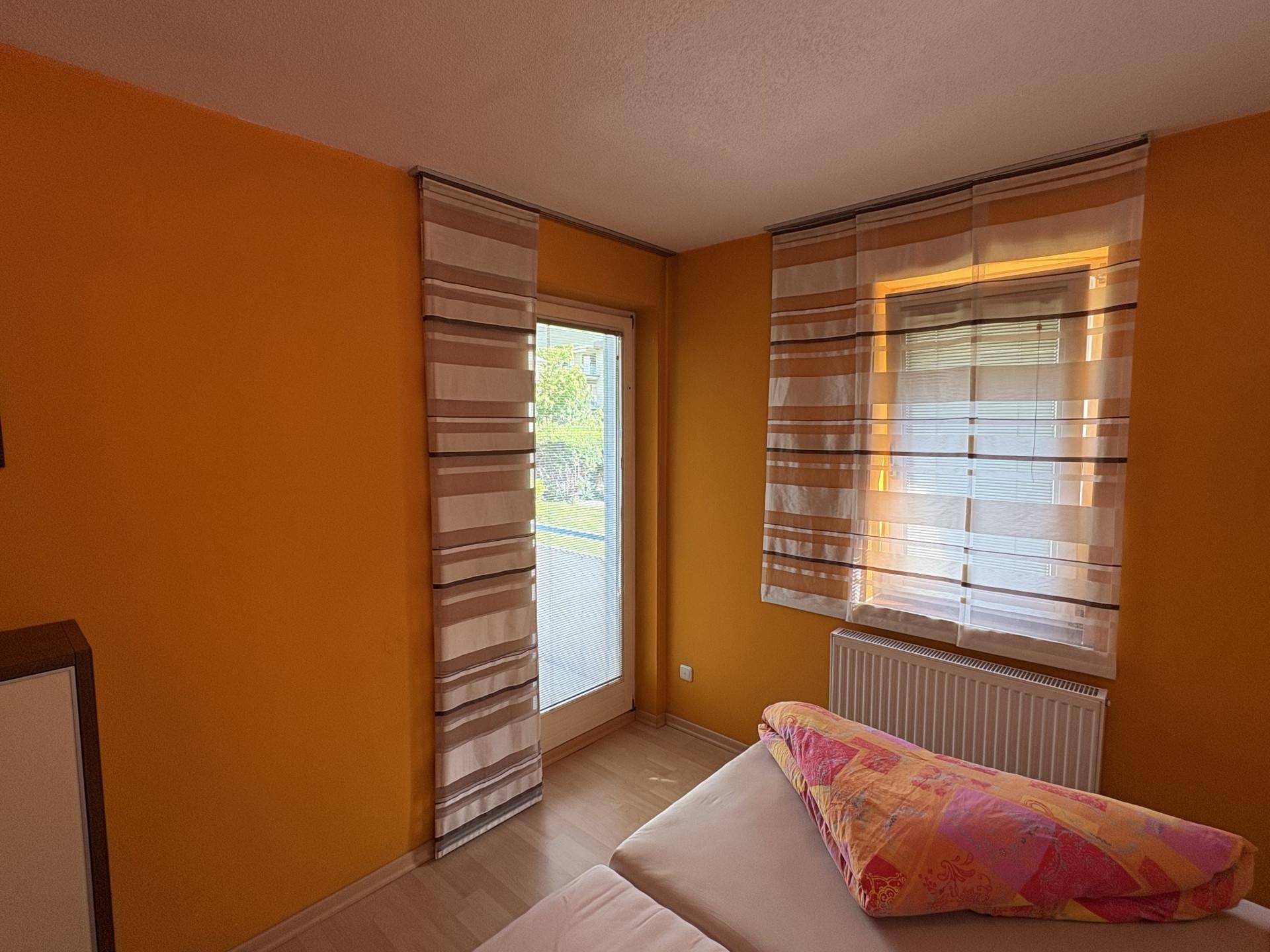 Helles Schlafzimmer mit Zugang zum Balkon und gelben Wänden, ausgestattet mit einem Heizkörper.
