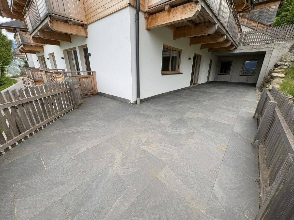 Große, geflieste Terrasse neben dem Gebäude mit Holzbalkonen und Blick ins Grüne.