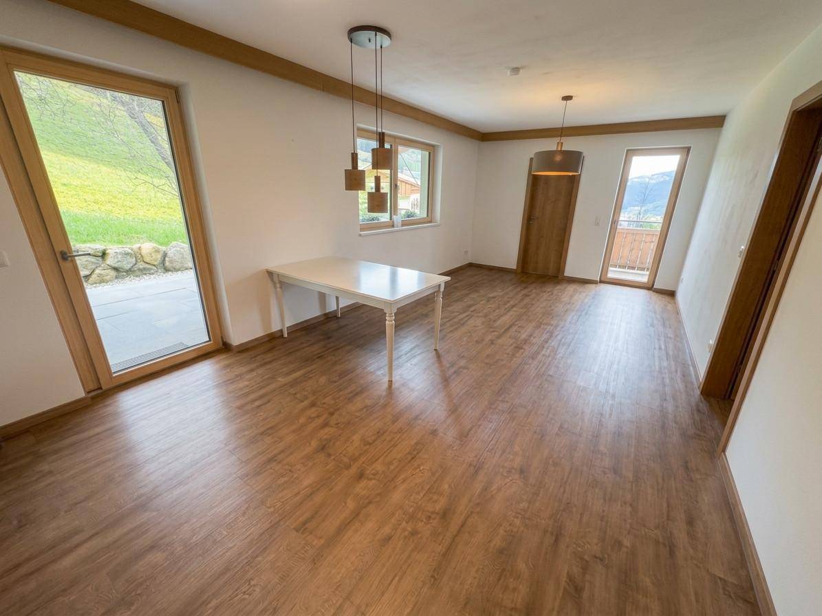 Helles Zimmer mit Holzboden, weißem Tisch und Zugang zu Terrasse und Balkon mit Bergblick.