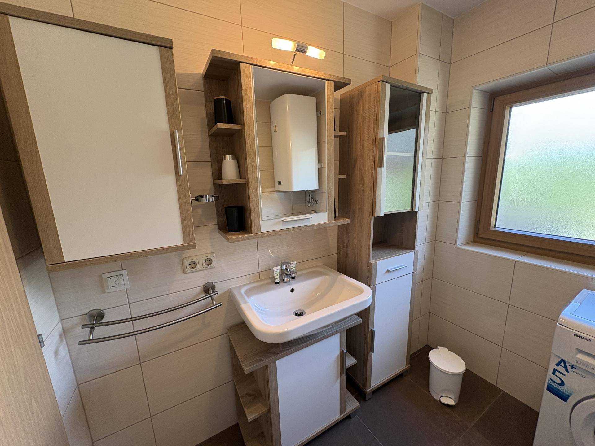 Modernes Badezimmer mit Waschtisch, Spiegelschrank, hellen Fliesen und einem Fenster.